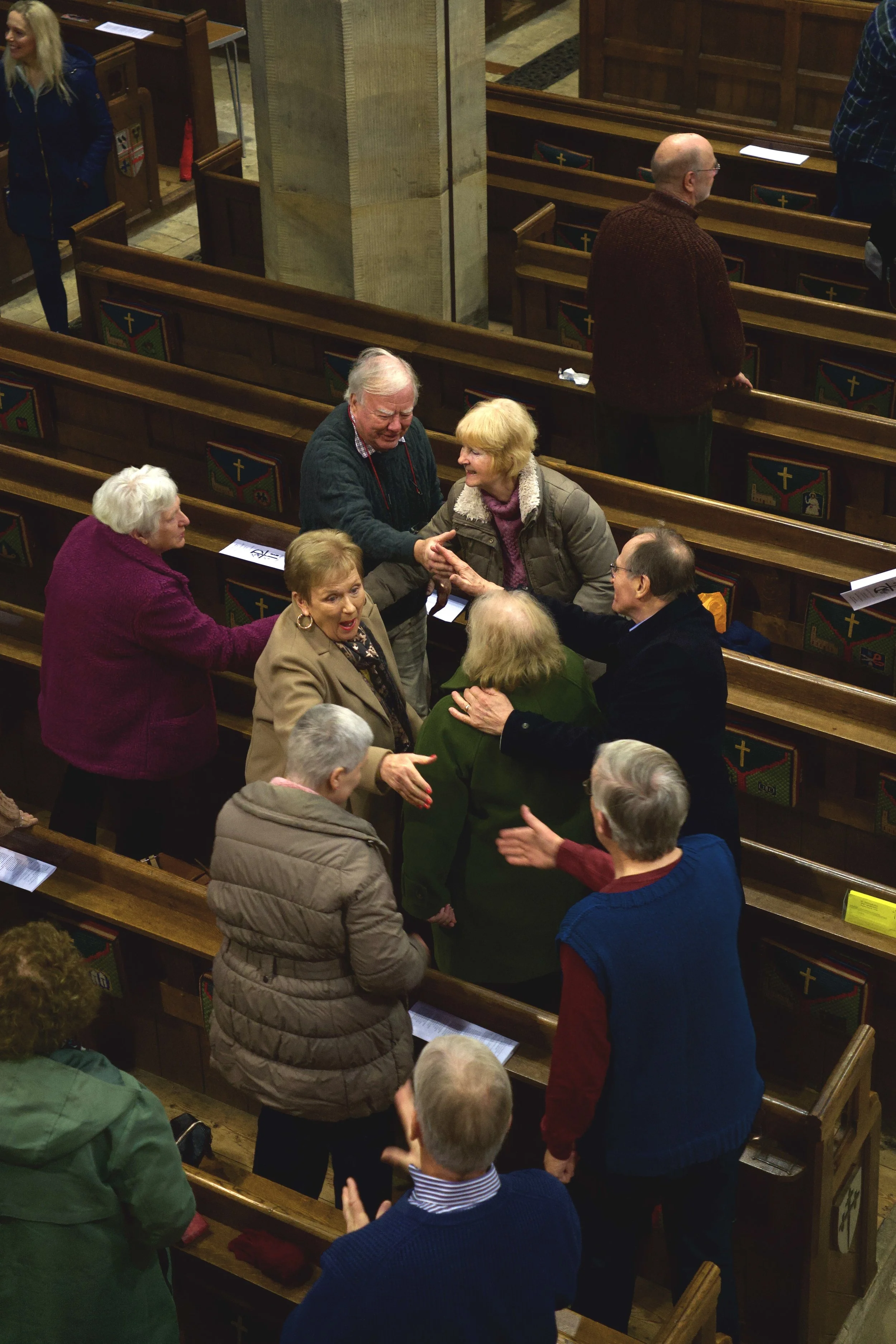 Group of elderly people greeting and shaking hands inside a church with wooden pews and religious symbols on the side.