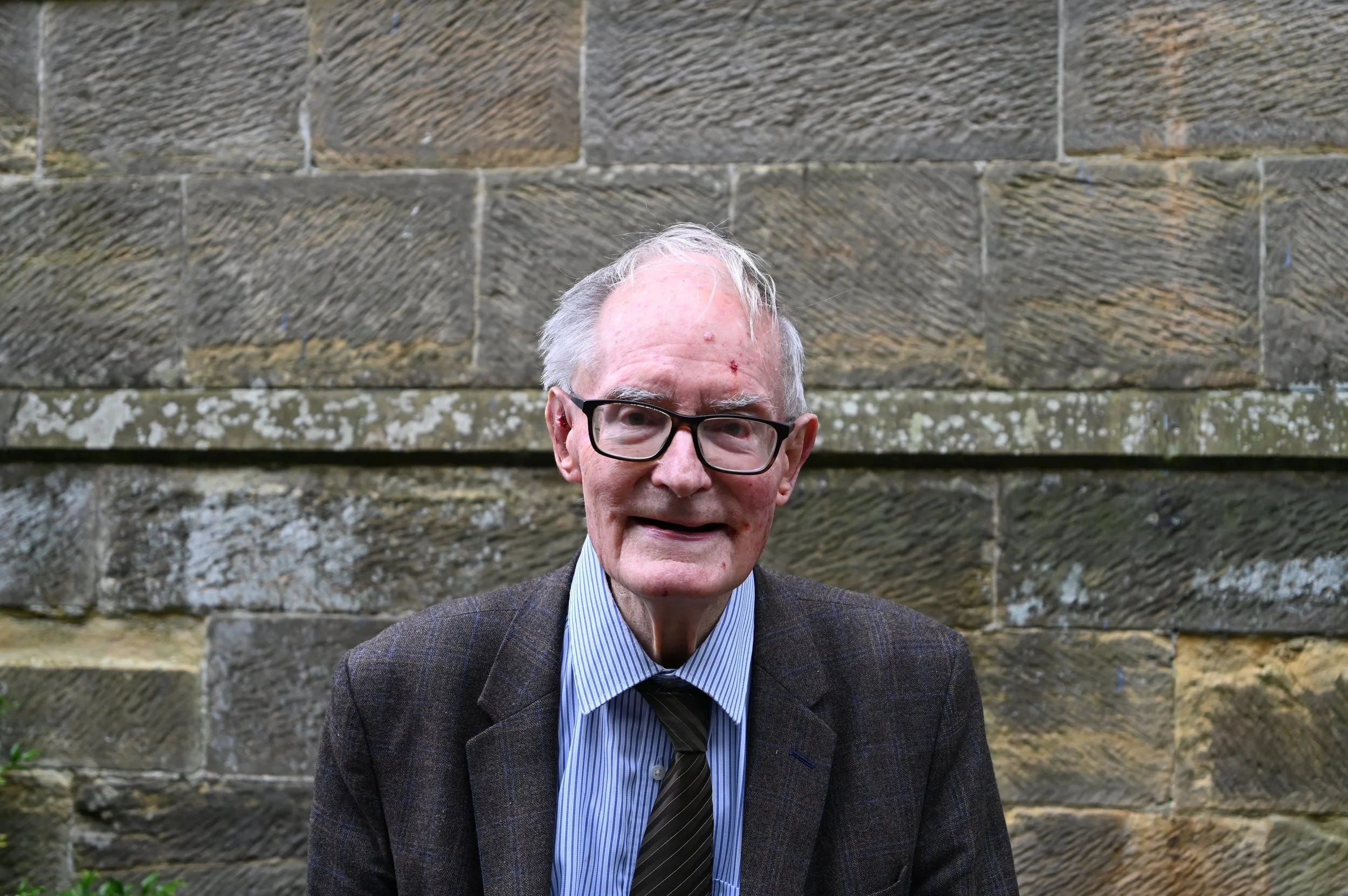 An elderly man with glasses, white hair, and a suit with a tie, standing outdoors against a stone wall.