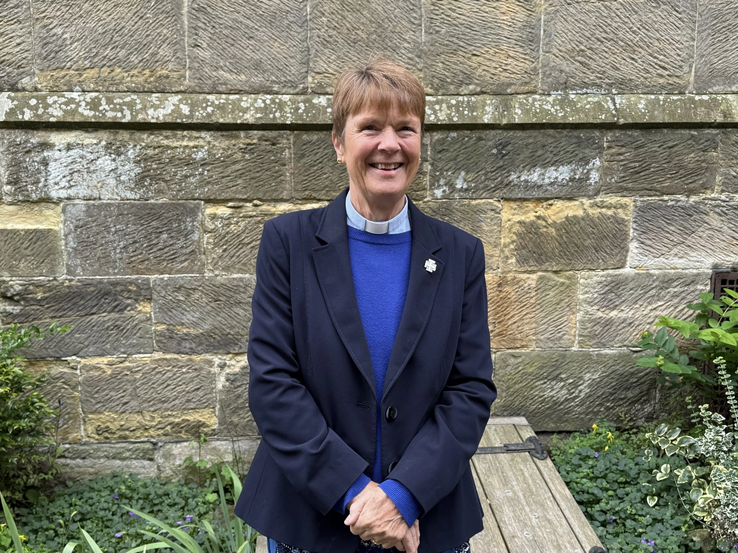 A smiling woman with short brown hair wearing a clerical collar, a blue sweater, and a dark blazer, standing outdoors in front of a textured stone wall and some greenery.