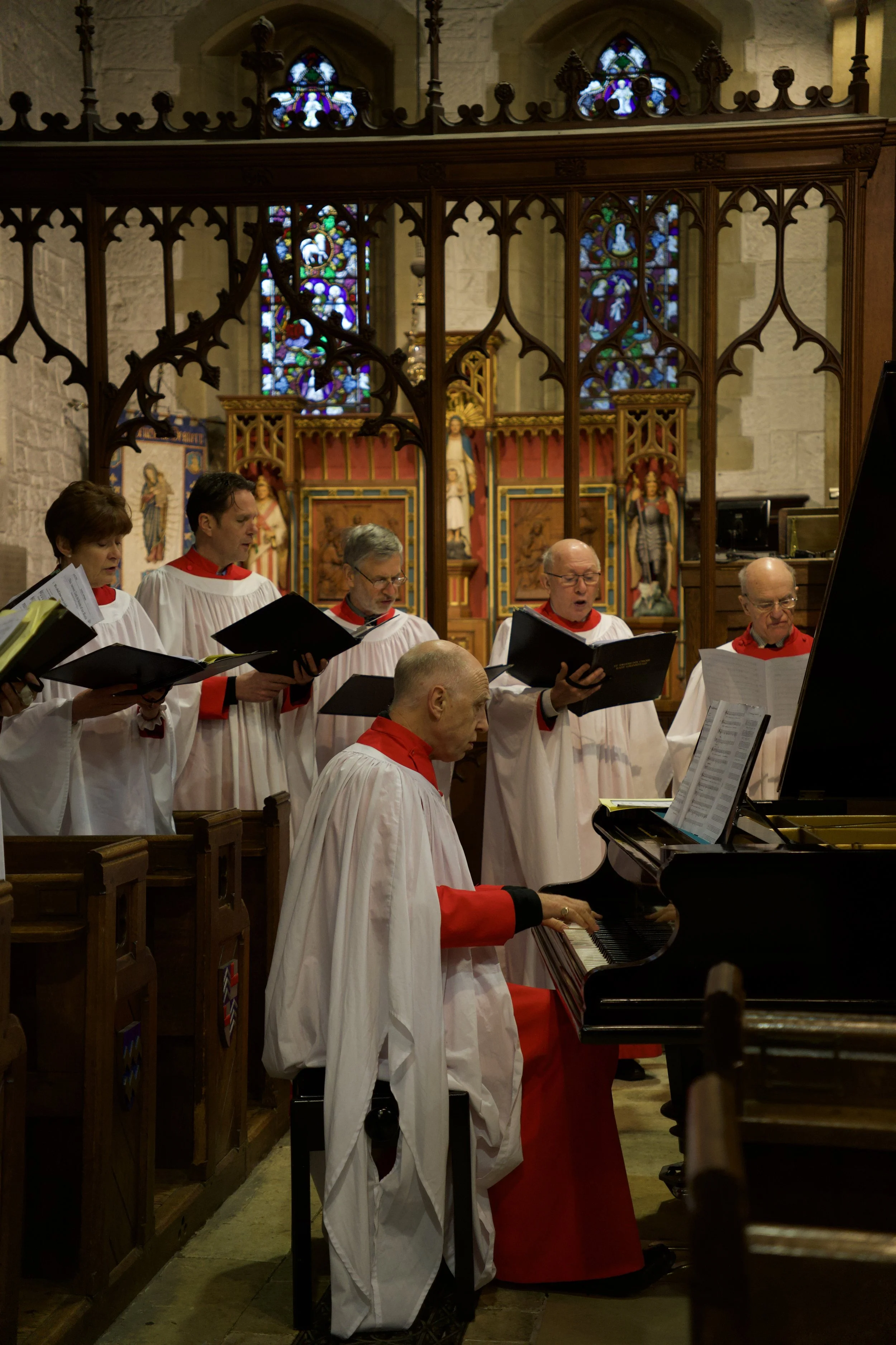 A choir of seven people, dressed in white robes with red accents, performing in a church behind a piano. The church has stained glass windows and religious statues in the background.