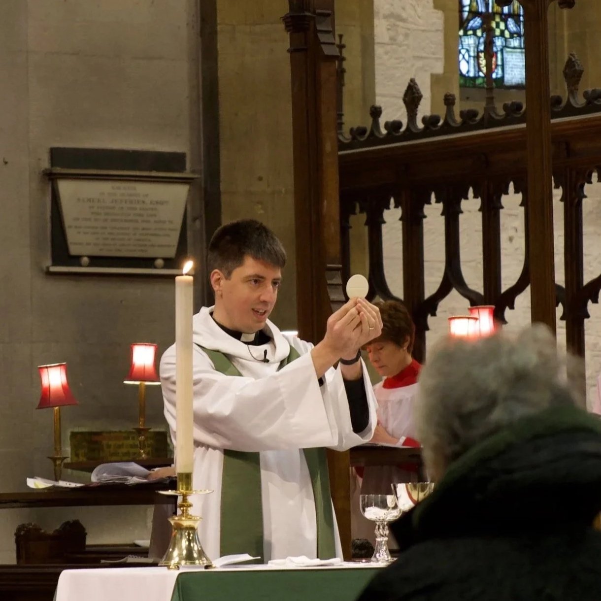 A priest in white robes holding a communion wafer during a religious service inside a church. The scene includes a table with lit candles, a chalice, and a congregant in the foreground, with stained glass windows in the background.