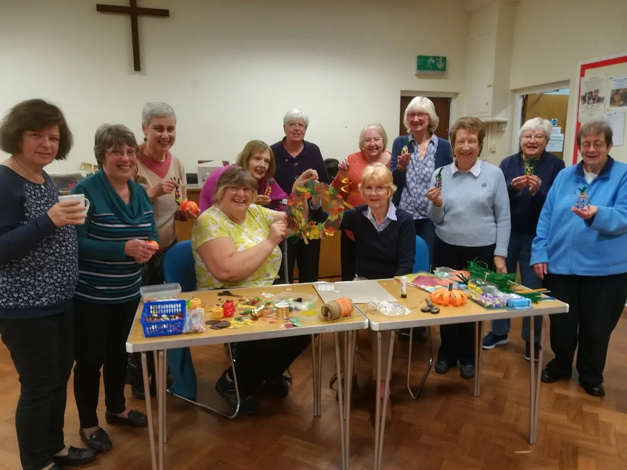 A group of women gathered around tables inside a church hall, holding decorated crafts and smiling, with a wooden cross on the wall in the background.