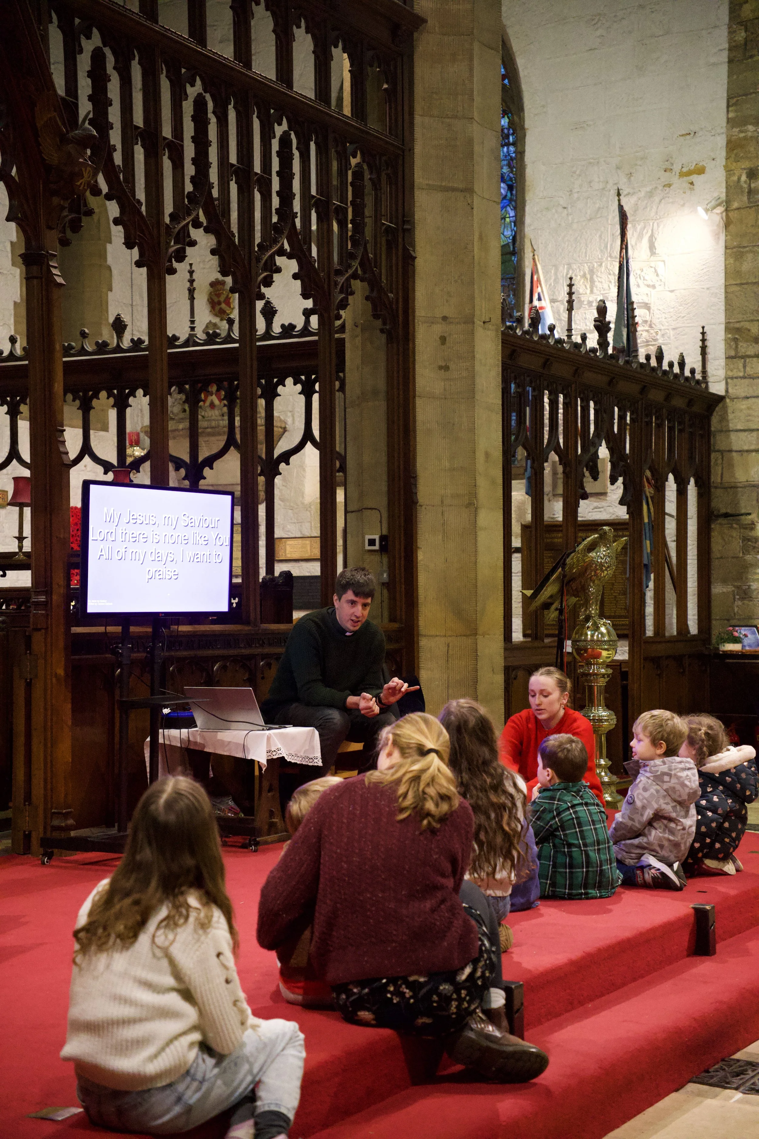 A man leading a religious lesson with young children seated on steps inside a church, with a screen displaying song lyrics and various flags and religious symbols visible.