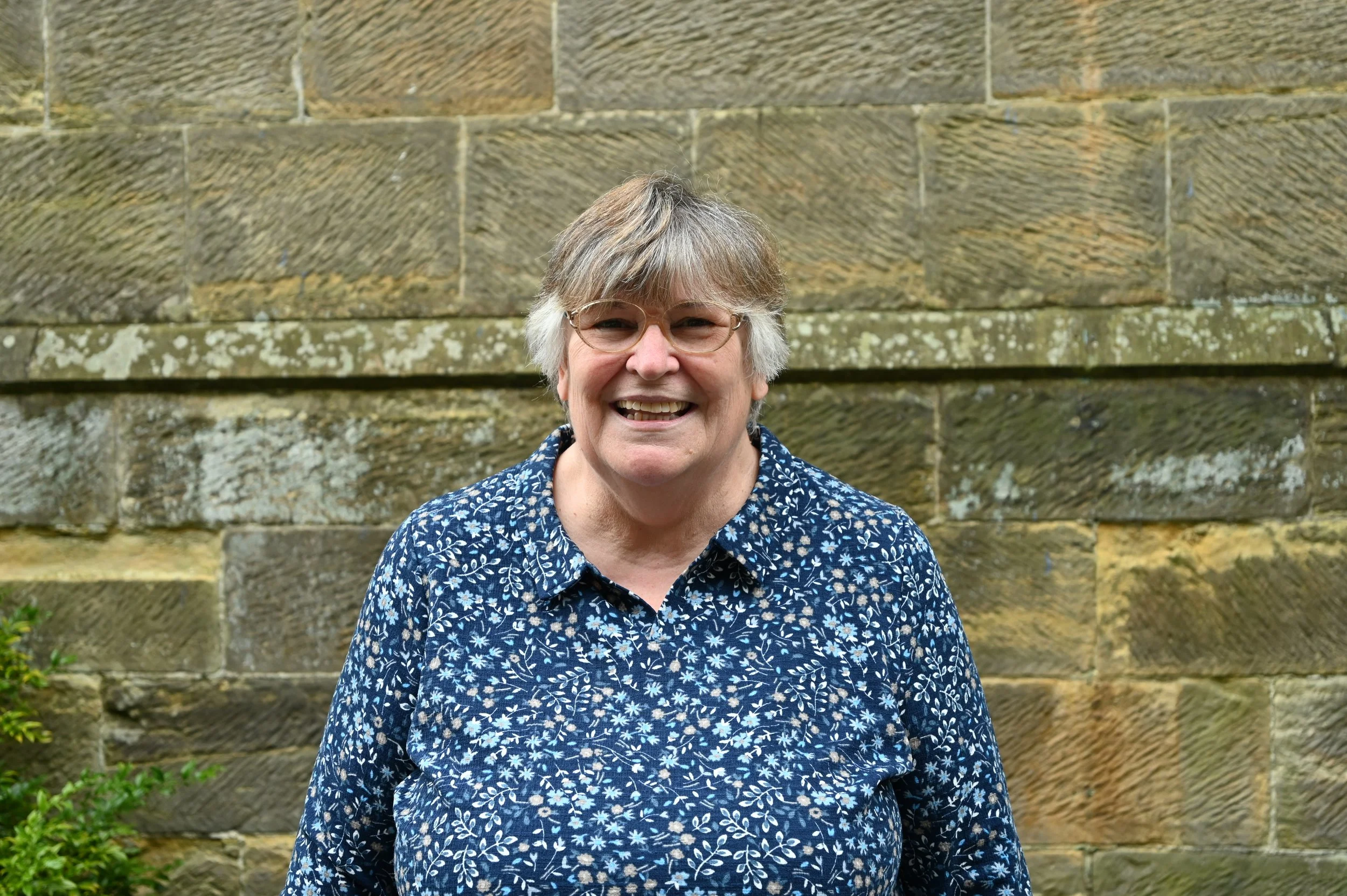 Smiling elderly woman with glasses, wearing a blue patterned shirt, standing in front of a stone wall.