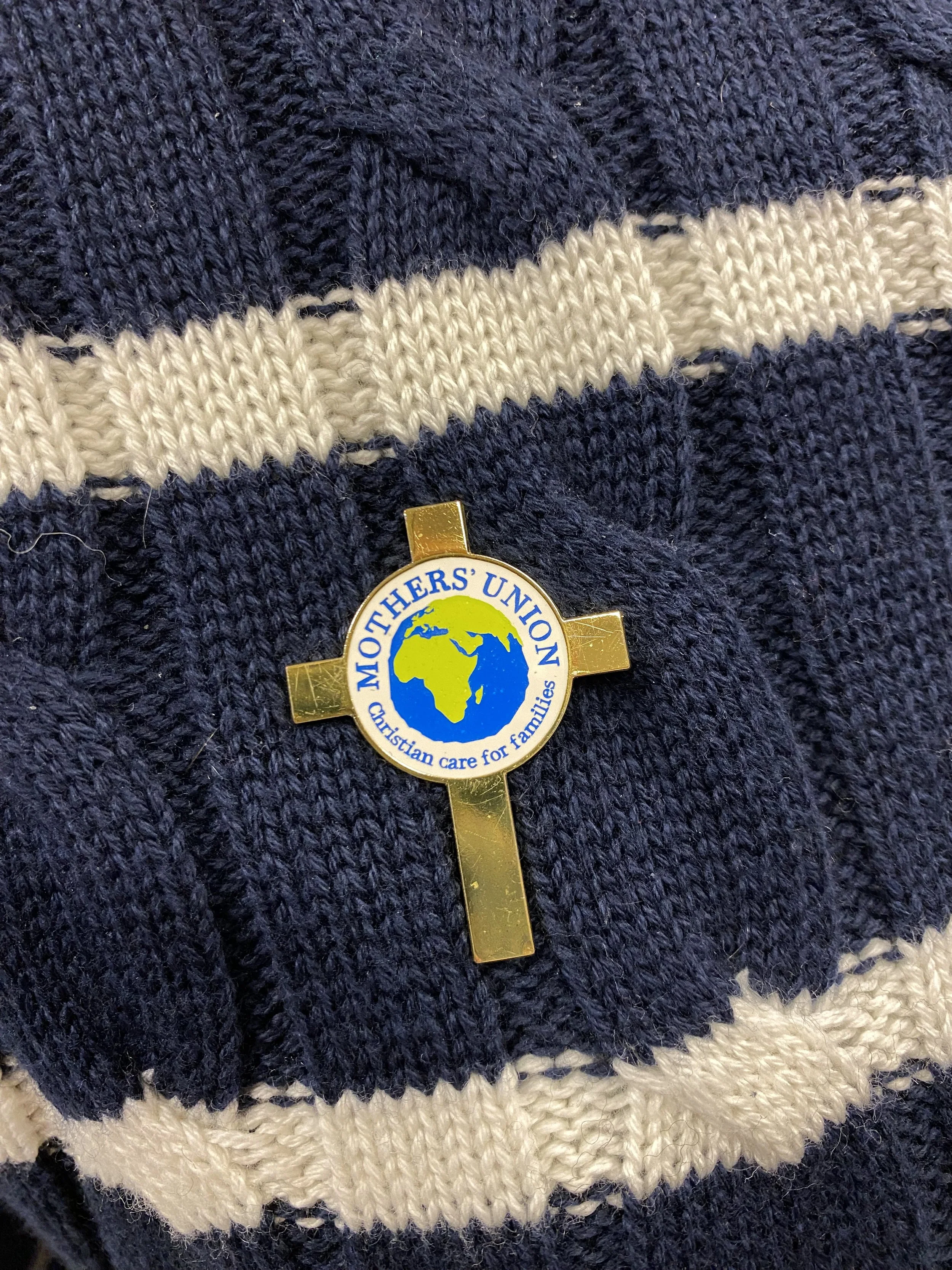 A gold cross pin with the logo of Mother’s Union, featuring a globe and the words 'Mother’s Union' and 'Christian care for families,' pinned on a dark blue and beige striped knitted fabric.