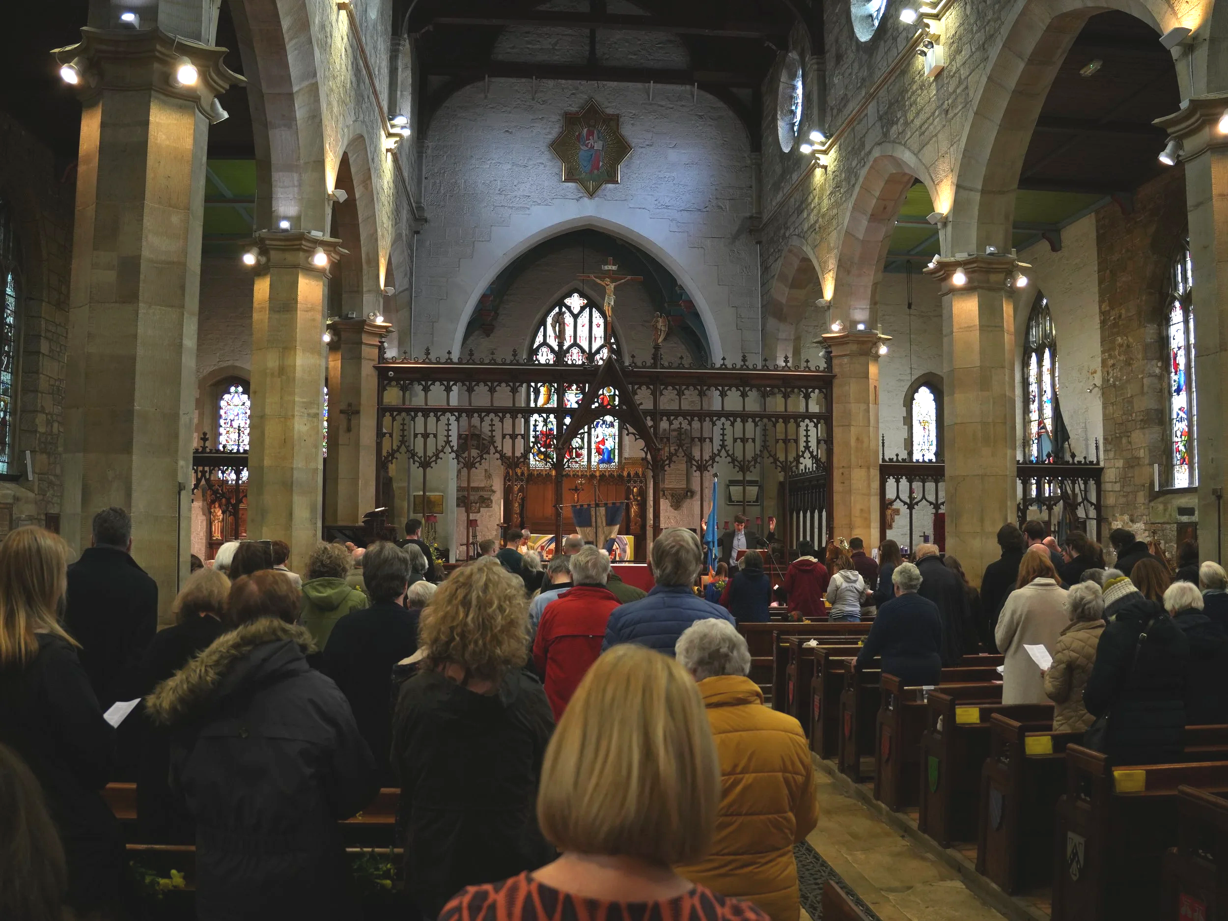 Inside a church during a service or event, with people gathered in pews facing the altar, which features a crucifix and religious statues. The church has stone walls, stained glass windows, and arched ceilings with warm lighting.