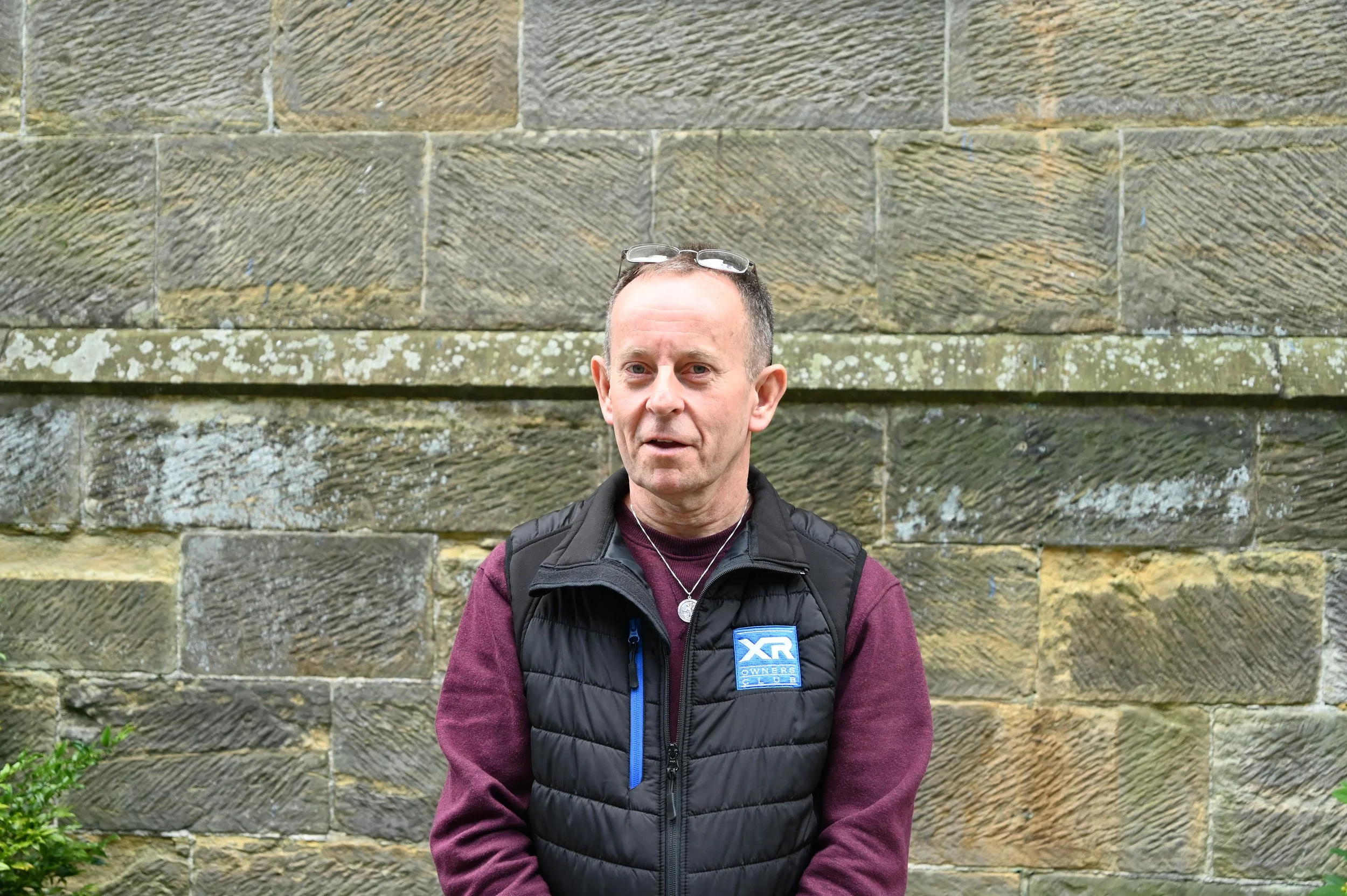 A middle-aged man standing outdoors in front of a stone wall, wearing glasses, a black padded vest with a blue logo, and a maroon long-sleeve shirt.