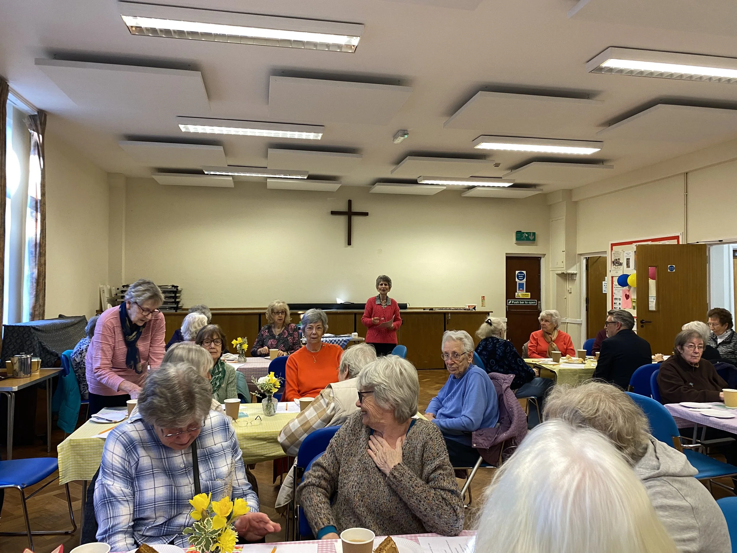 A group of elderly people gathered in a community hall. Some are sitting at tables with yellow tablecloths, drinking from paper cups, and talking. There is a woman standing in the middle of the room addressing the group. The room has a crucifix on the wall above a stage area, and there are balloons and decorations on the walls.