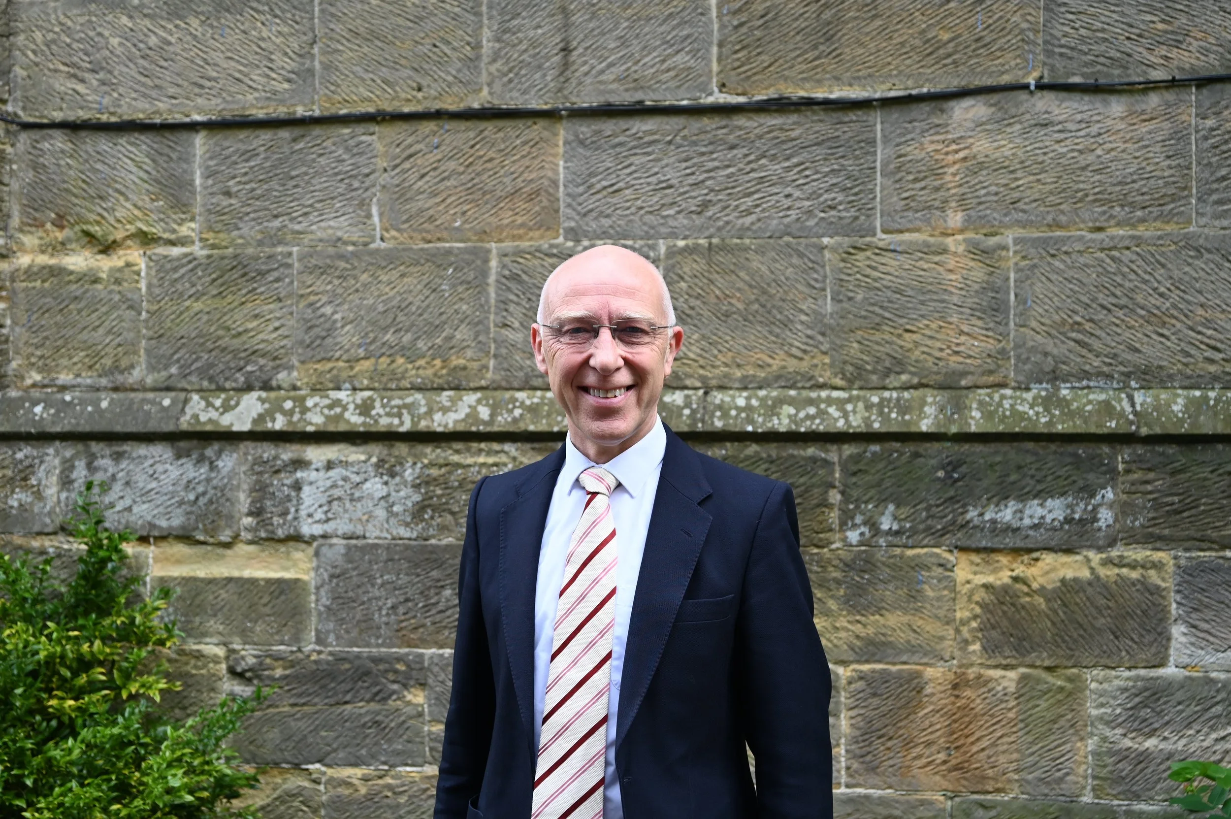 A smiling man dressed in a suit and tie standing outdoors in front of a stone wall.