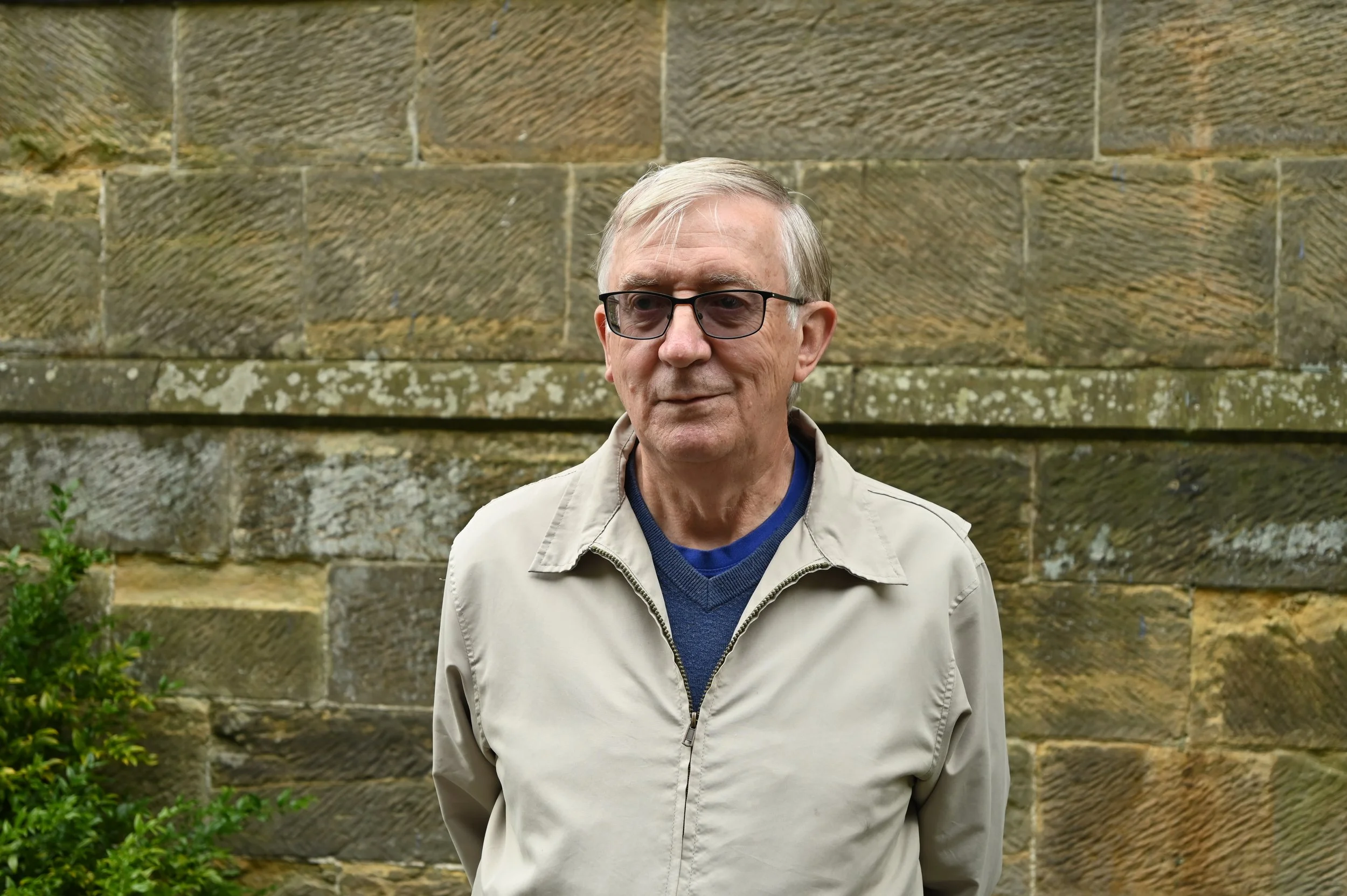Older man with glasses and light-colored jacket standing outdoors in front of a stone wall.