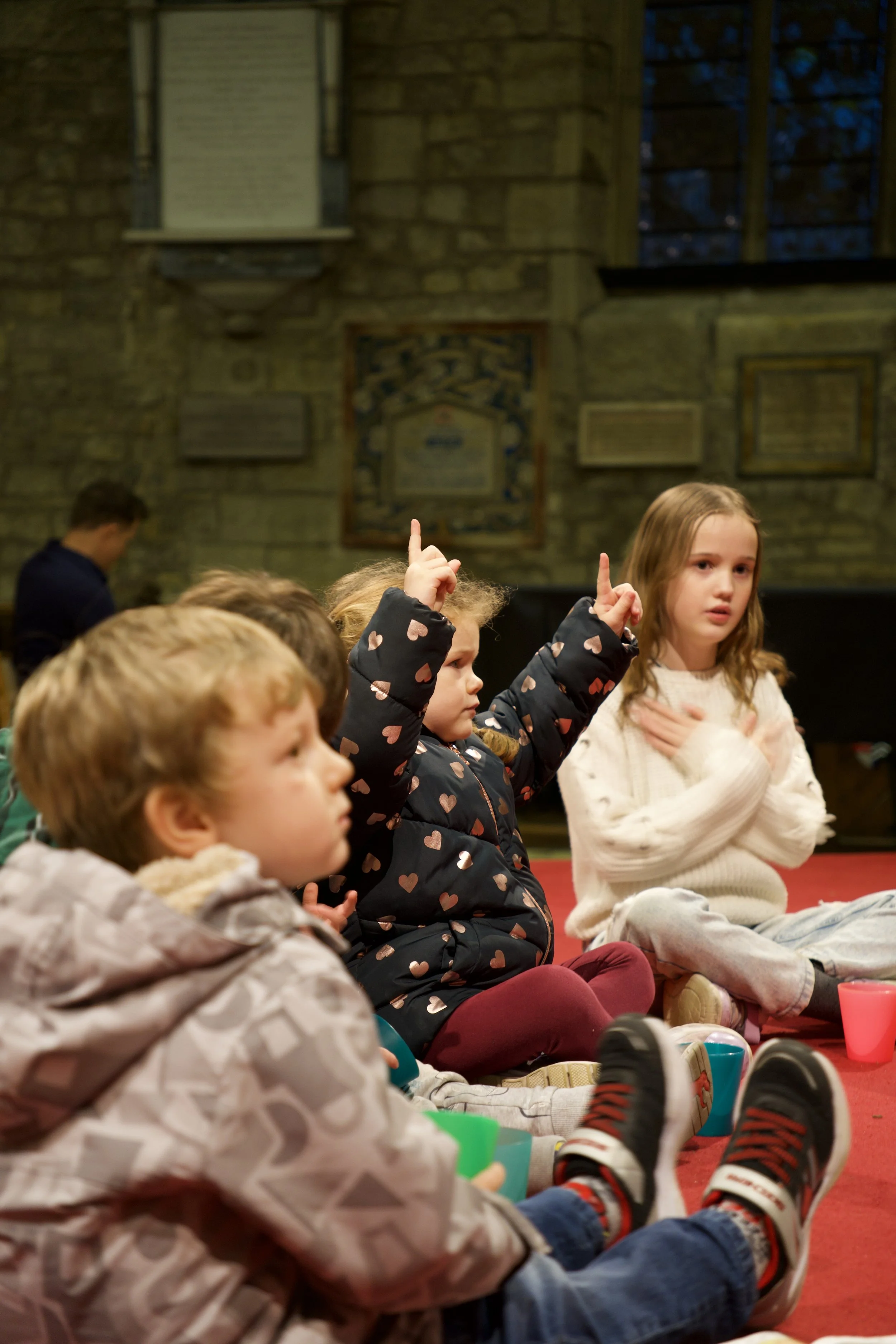 Children sitting on the floor during a group activity or lesson inside a building with brick walls and framed artwork.