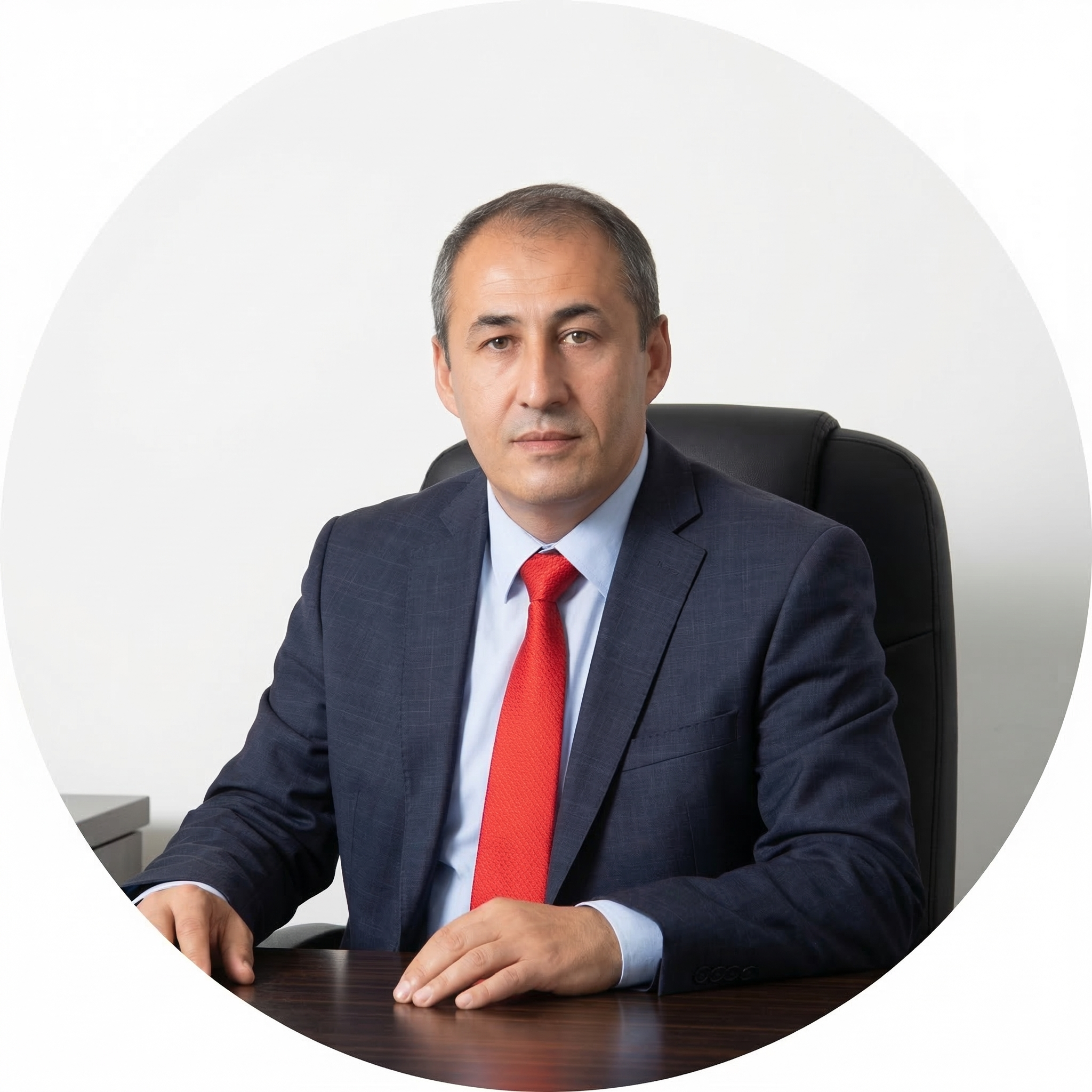 Ofis ortamında, sandalye arkası ve masanın üzerinde oturan erkek, ciddi ifadeyle kamera bakıyor, lacivert takım ve kırmızı kravat giyiyor.