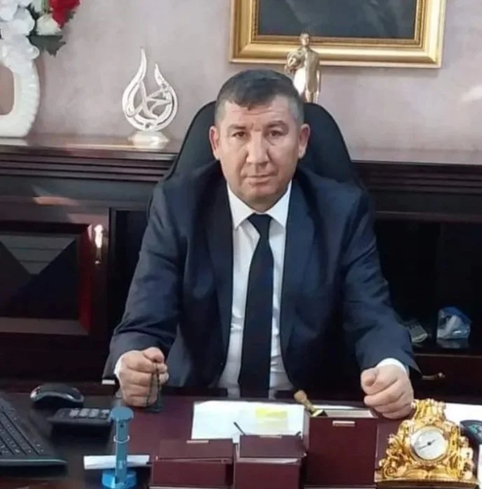 Ofis ortamında, masa başında siyah ceket ve bağlamalı gömlek giymiş, oturan bir erkek. Masasında çeşitli ofis malzemeleri ve lüks bir altın detaylı saat var, arka planda çerçeve içinde tablo ve dekoratif objeler yer alıyor.