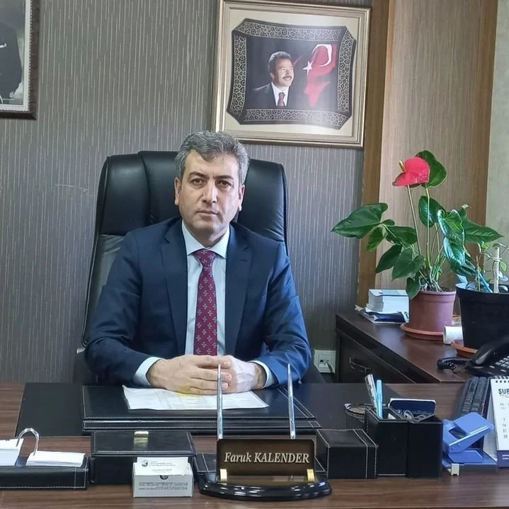 Ofis ortamında, masasında duran ve üzerinde 'Faruk KALENDER' yazan isim plakası olan erkek yönetici. Arka planda, duvarda, Türkiye ve Cumhurbaşkanı'nın resmi. Yanında bir bitki var.