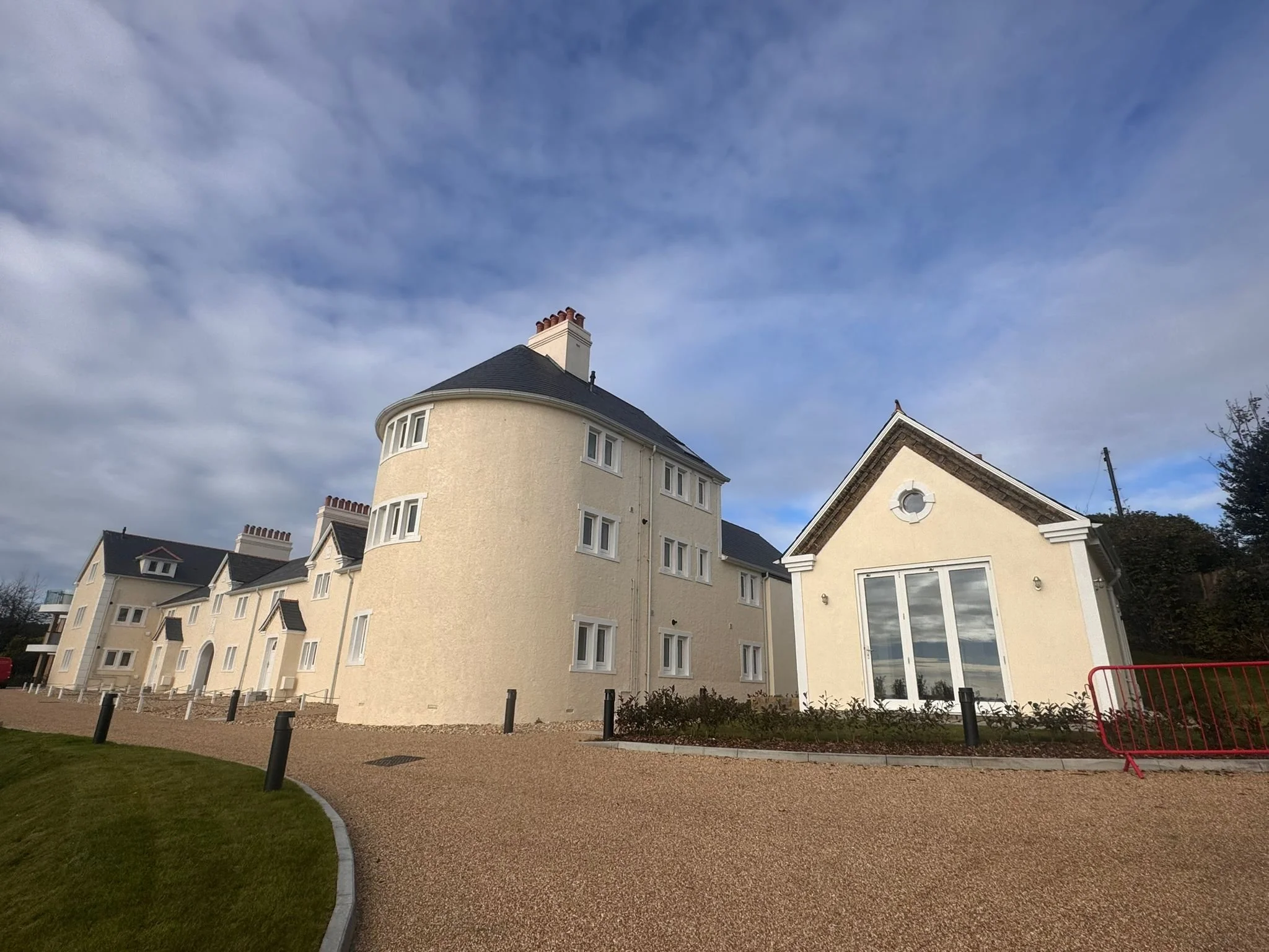 A large multi-story house with a rounded turret section, cream-colored exterior, many windows, and a dark gray roof. It is set against a partly cloudy sky. There is a gravel path bordered by grass and a row of short post bollards in front of the hous