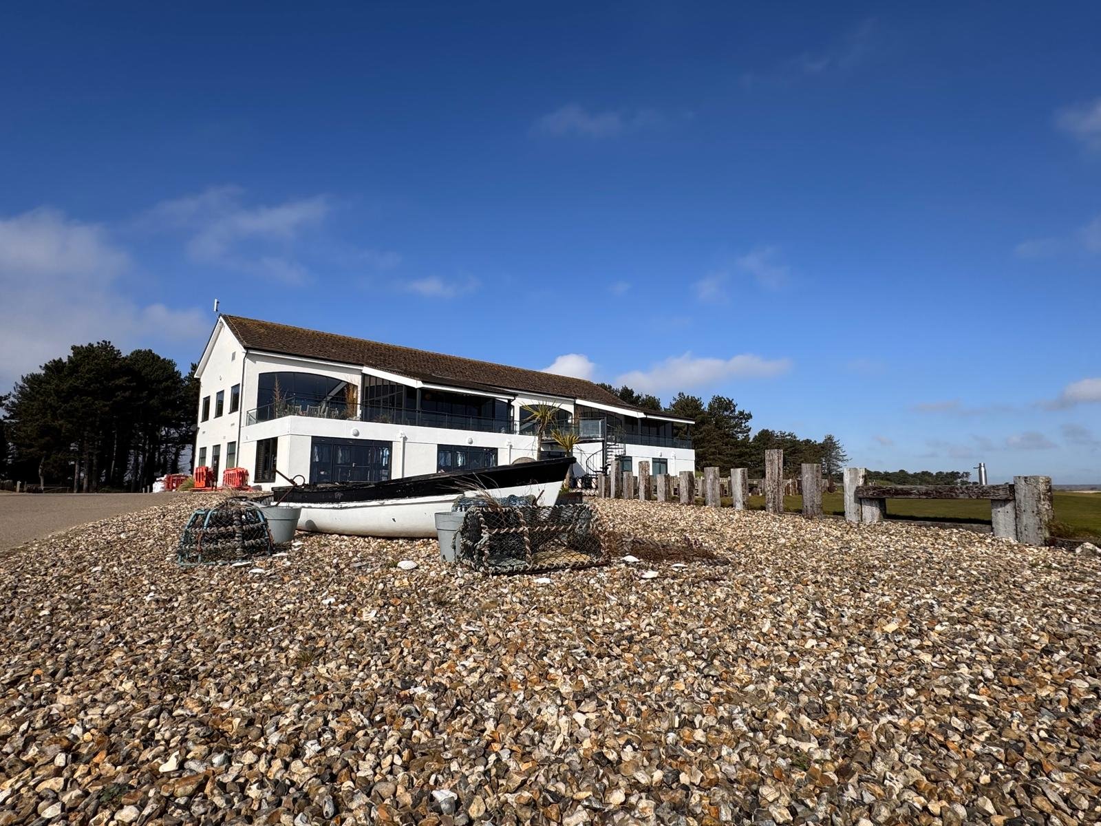A white house with large glass windows on a pebbled shoreline, a small boat resting on the pebbles, and a line of wooden posts with a clear blue sky overhead.