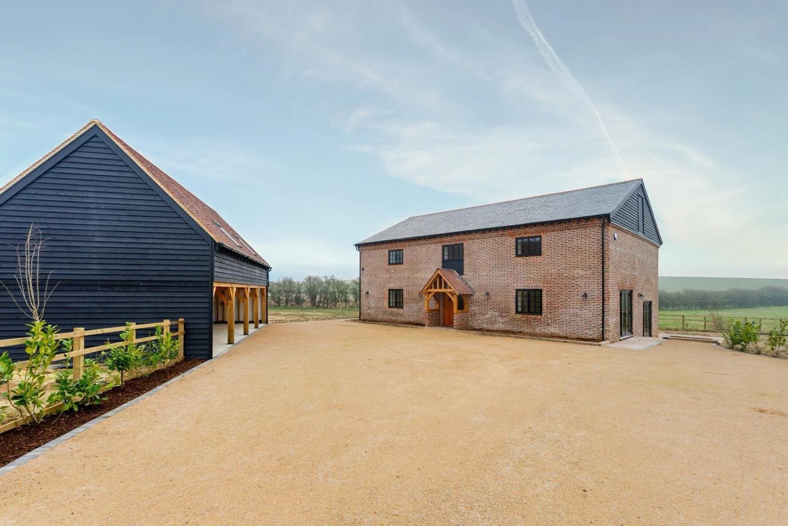 A modern brick house with black-framed windows and a small porch, and an adjacent black wooden barn with an open side, set on a gravel driveway with small plants along the edges, in a rural landscape under a partly cloudy sky.
