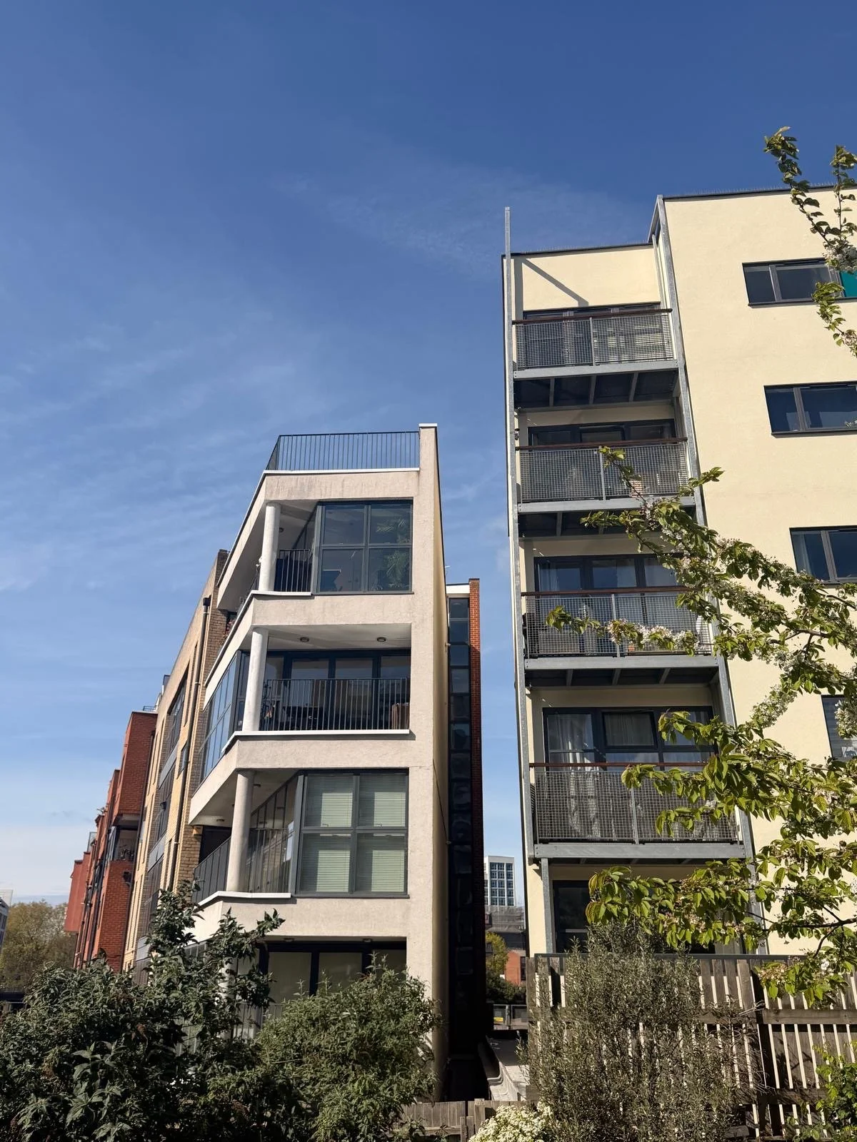 Modern multi-story apartment buildings with large windows and balconies, set against a clear blue sky, with trees and greenery in the foreground.