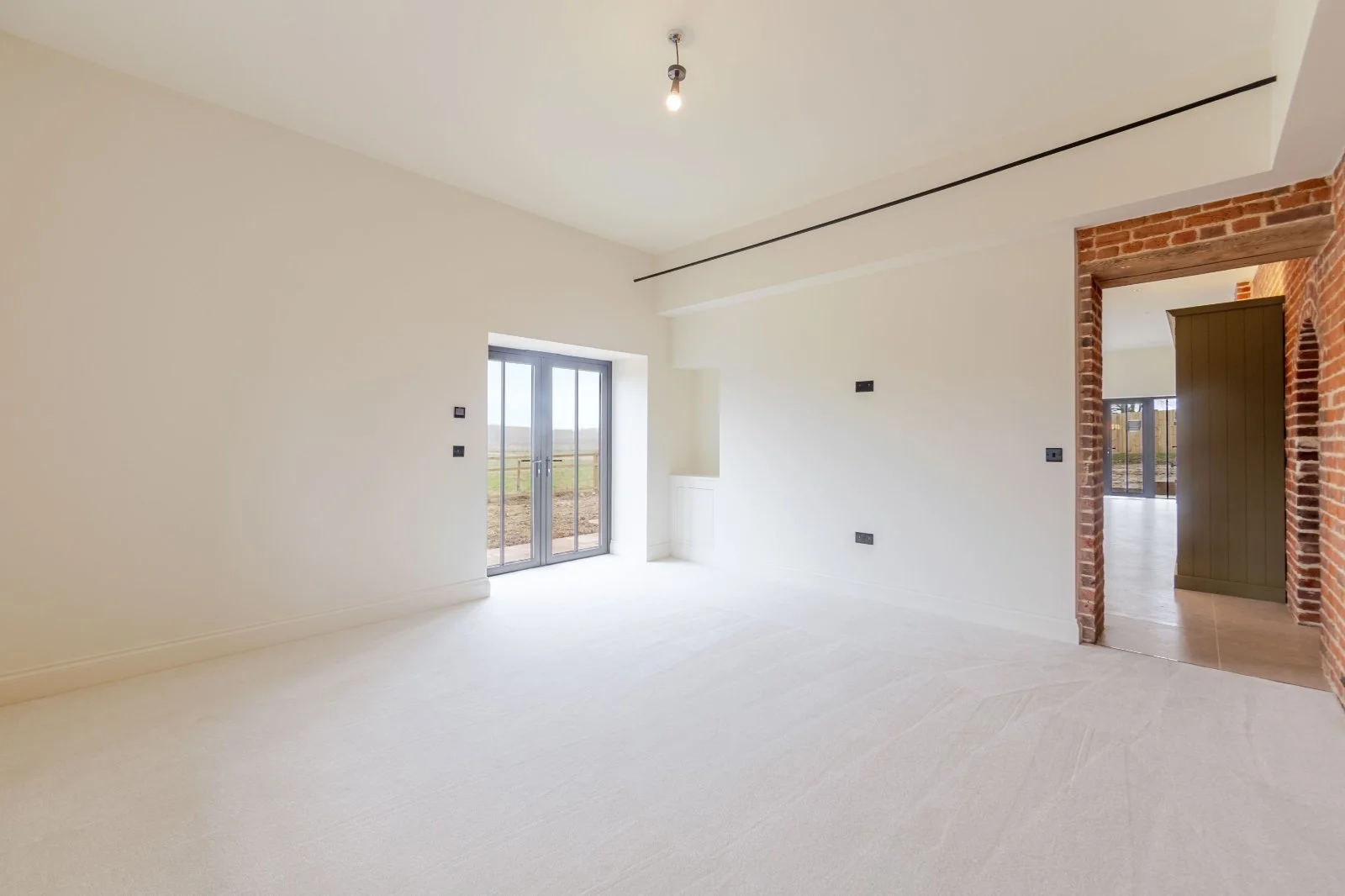 Empty room with white walls, large glass sliding doors, and a doorway with exposed red brick wall leading to another room.