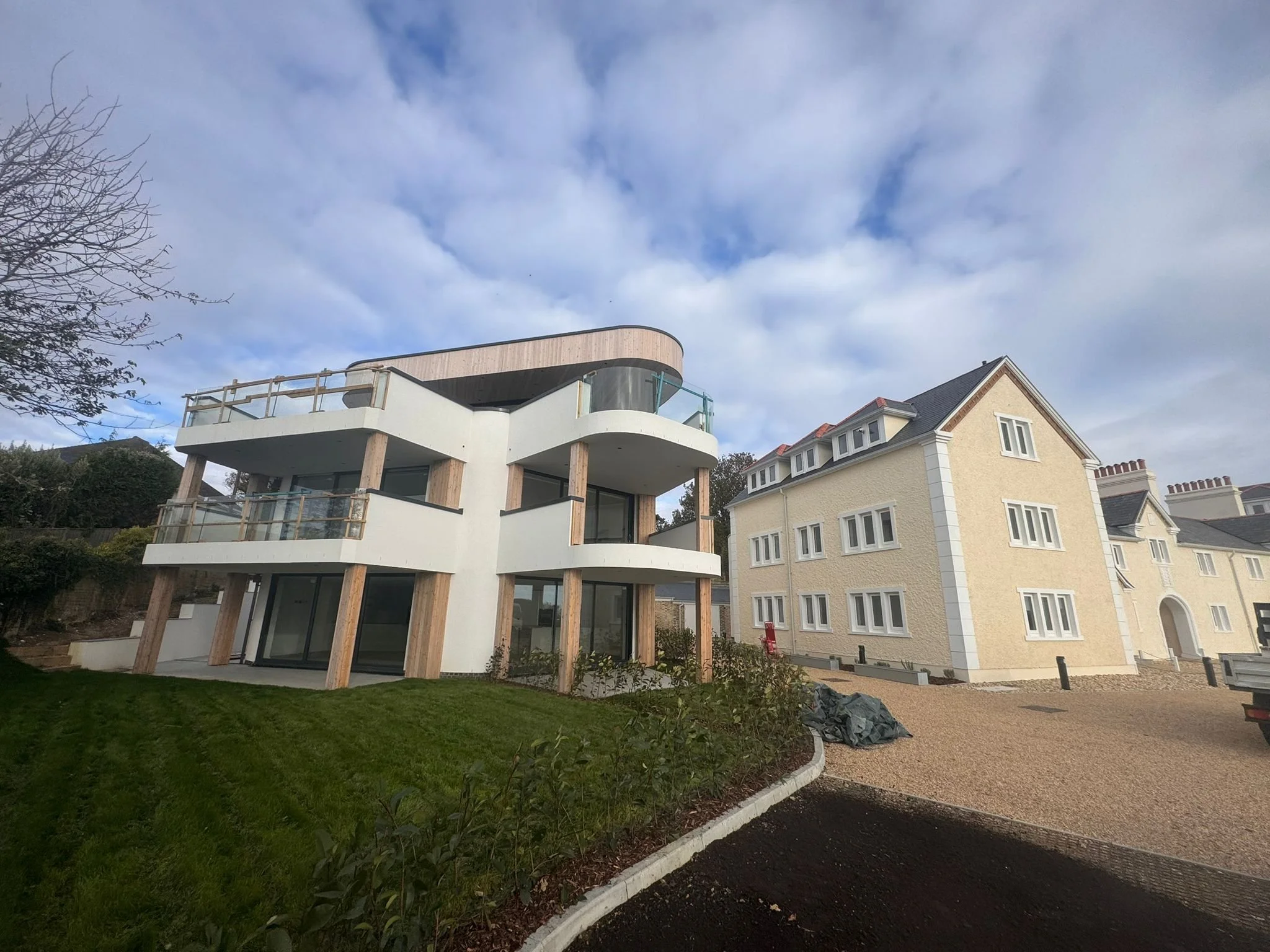 Modern house under construction with a curved balcony, white exterior, and wooden support beams, next to traditional yellow house with white trim, on a partly cloudy day.