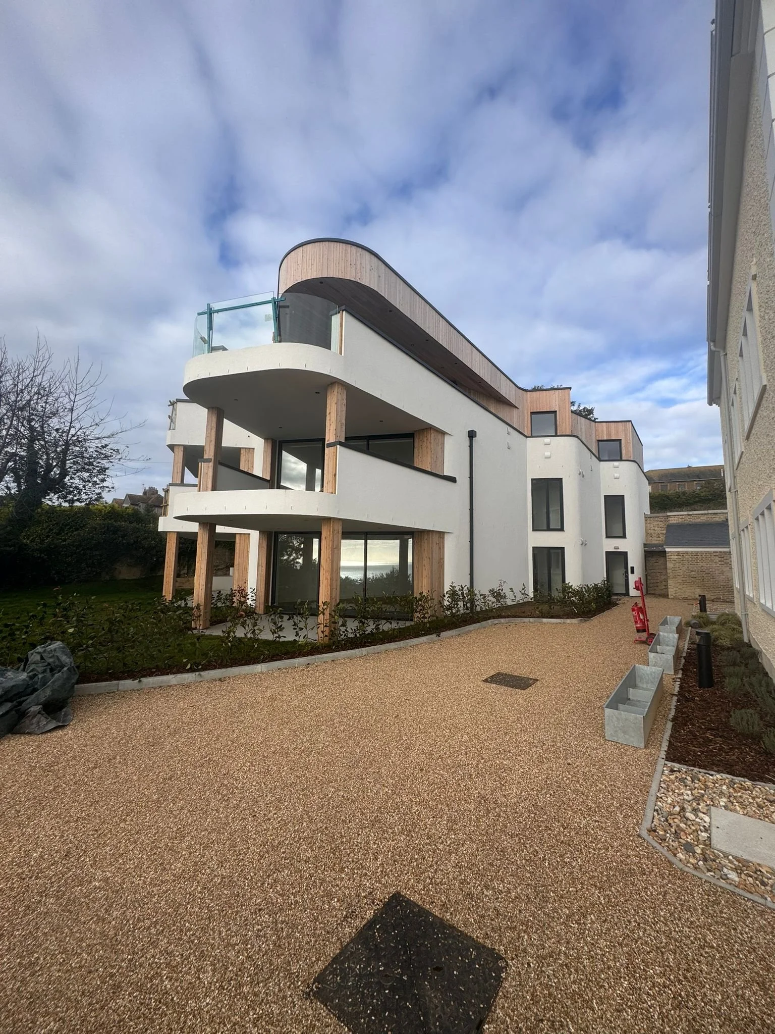 Modern multi-story building with curved balconies, wooden accents, and glass railing, surrounded by a landscaped area with a gravel walkway and planters.