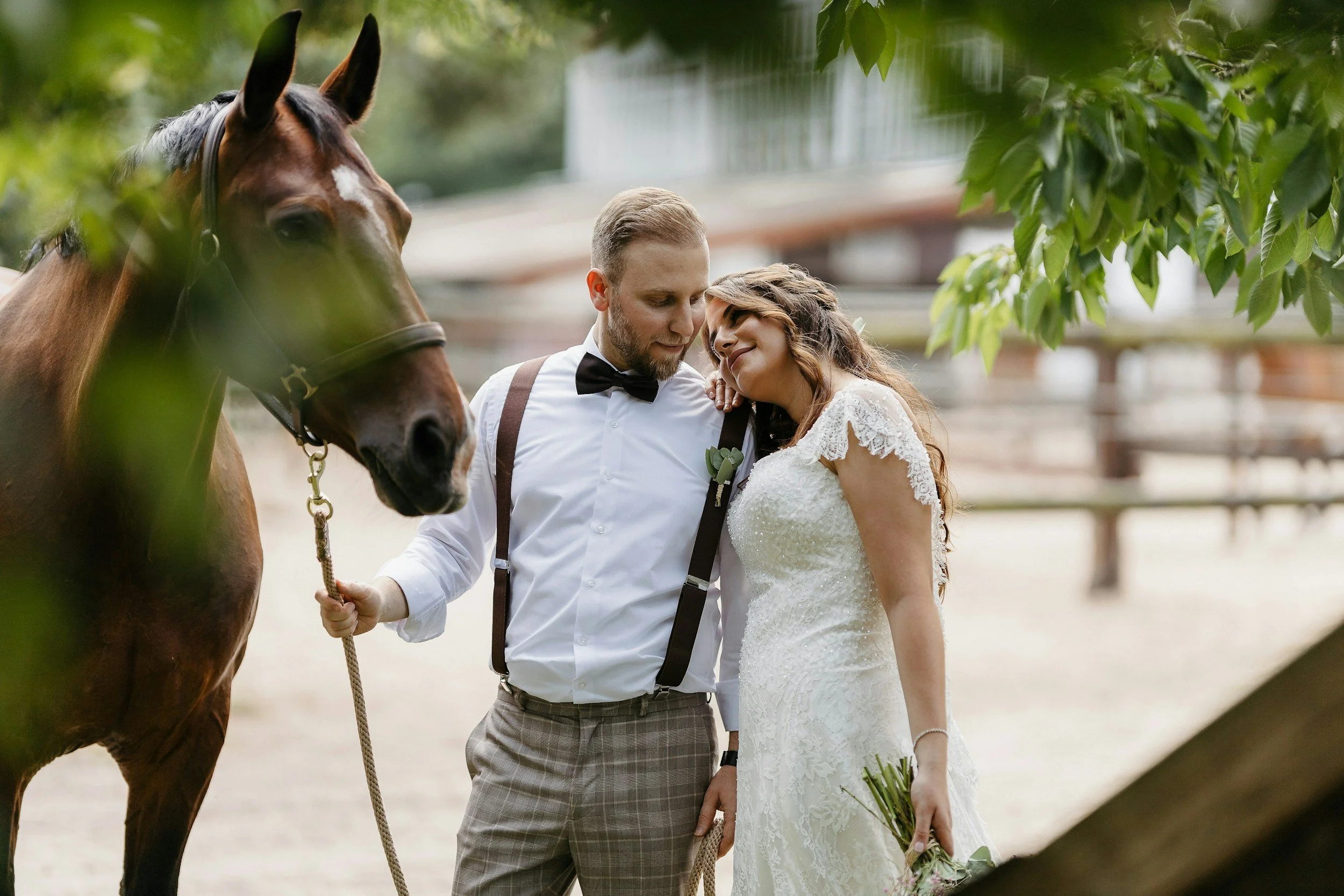 A bride and groom in wedding attire standing close together near a brown horse, with the bride holding a bouquet of flowers and leaning on the groom, in an outdoor setting with trees and a wooden fence.