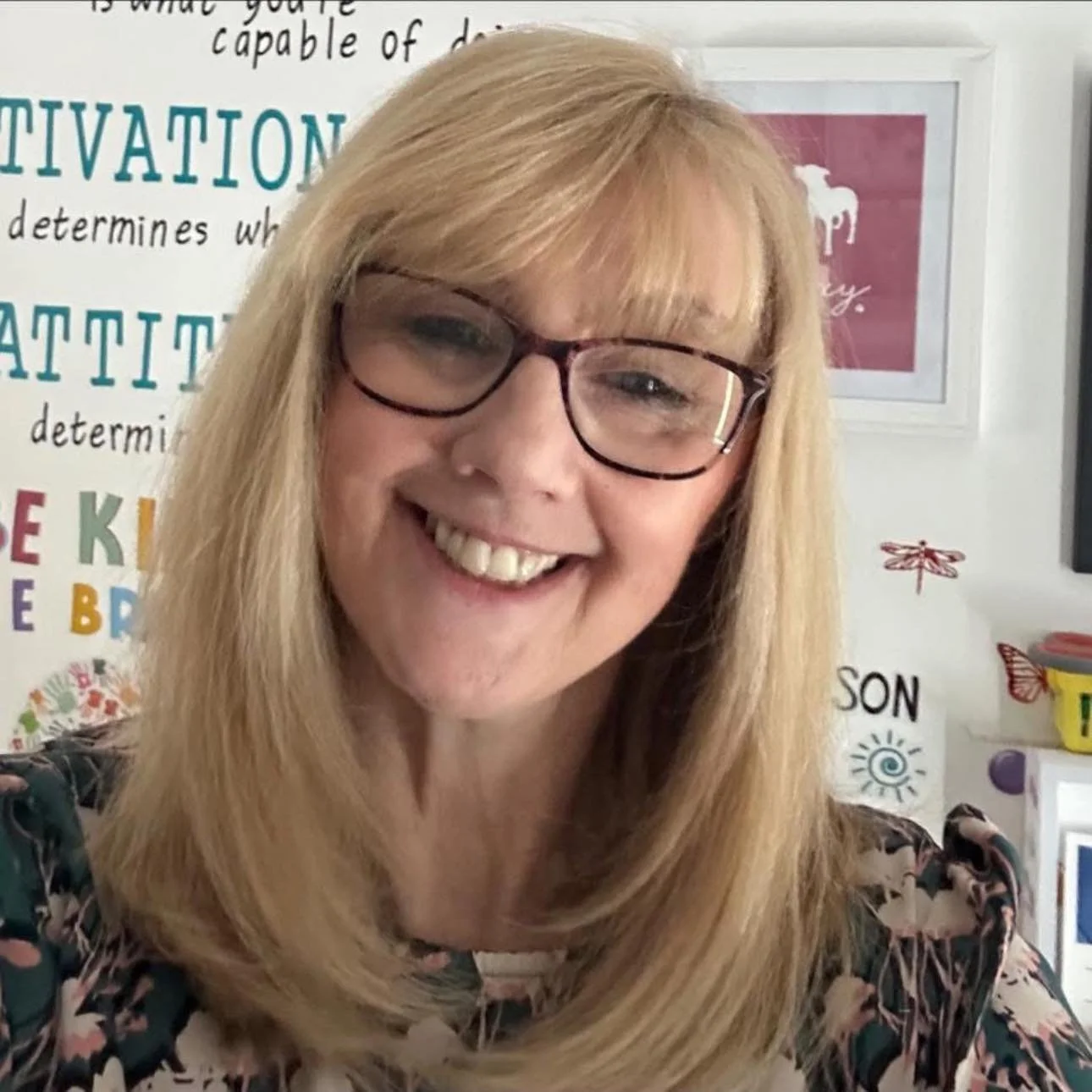 A woman with blonde hair and glasses smiling for a selfie in front of a colorful classroom wall.