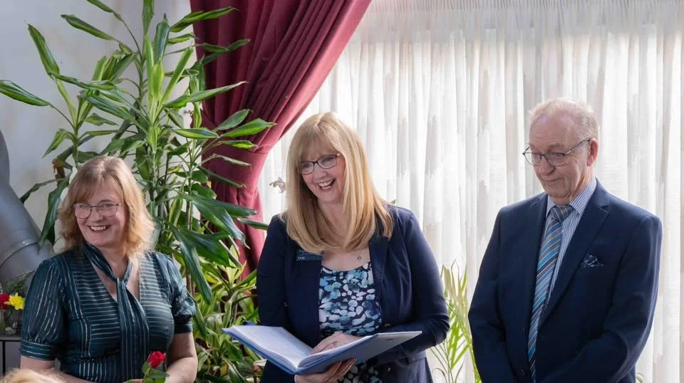 Three people, two women and one man, standing indoors near large potted plants and curtains, smiling and engaged in conversation.