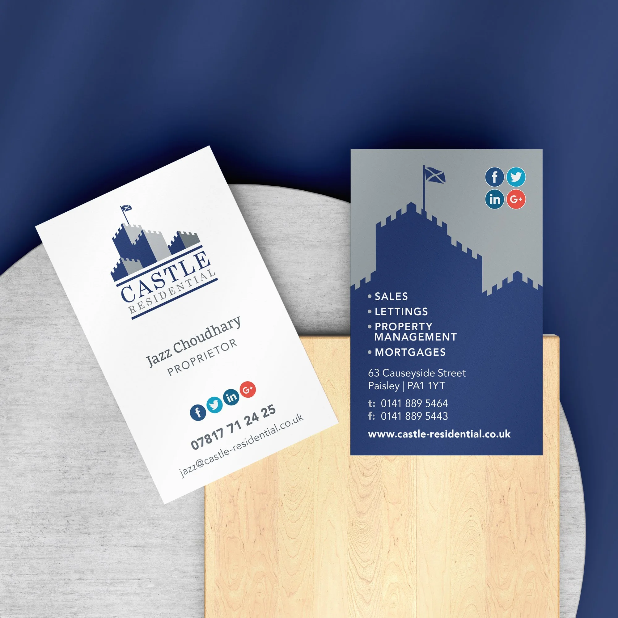 Two business cards for Castle Residential, one white for Jazz Choudhary with contact info and social media icons, the other dark blue with property services details and social media icons, placed on a light gray and a light wood surface.