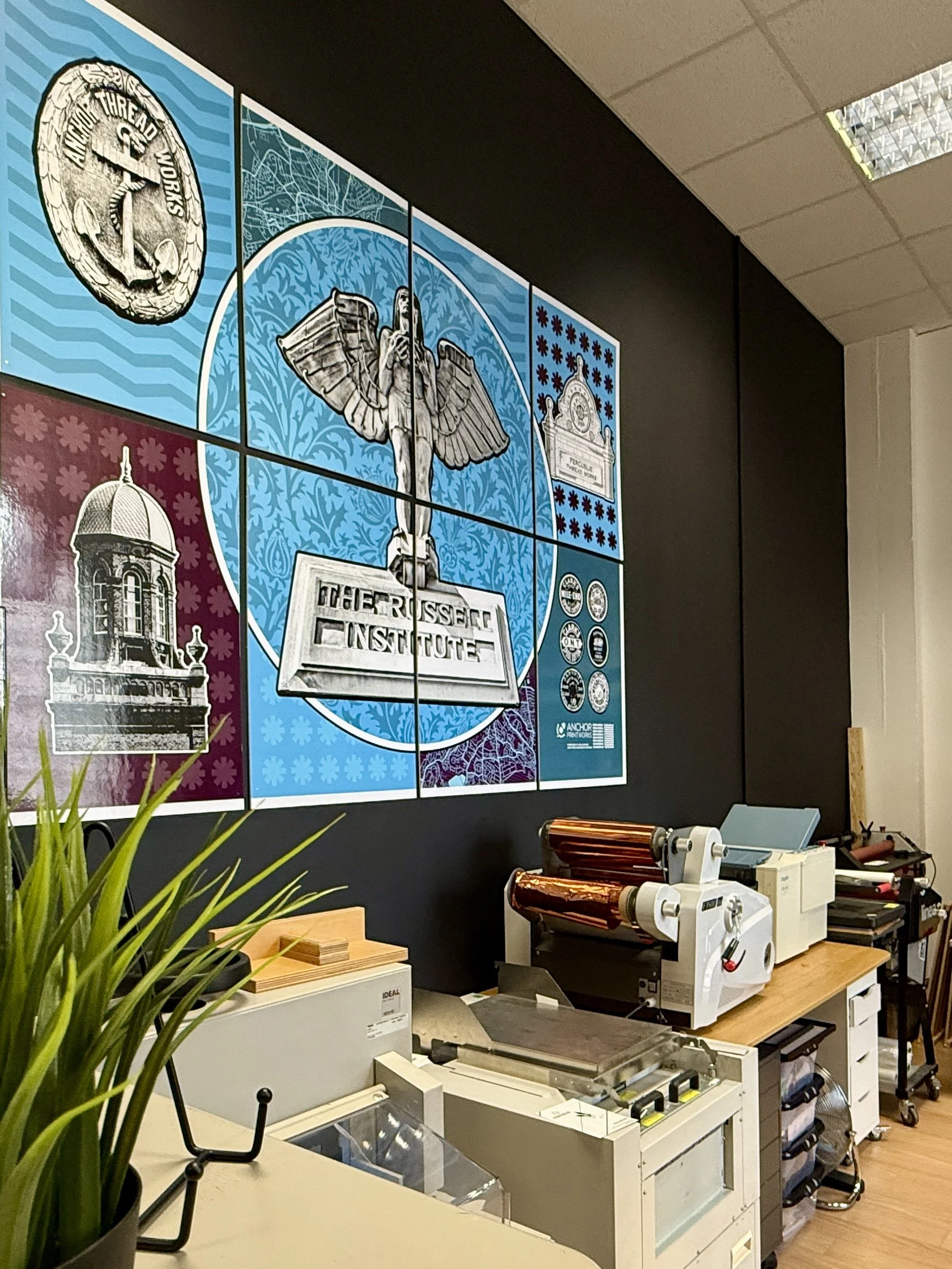 Office wall with a large multi-panel poster featuring logos and artwork related to The Russel Institute, with printing or packaging machines on a nearby countertop.