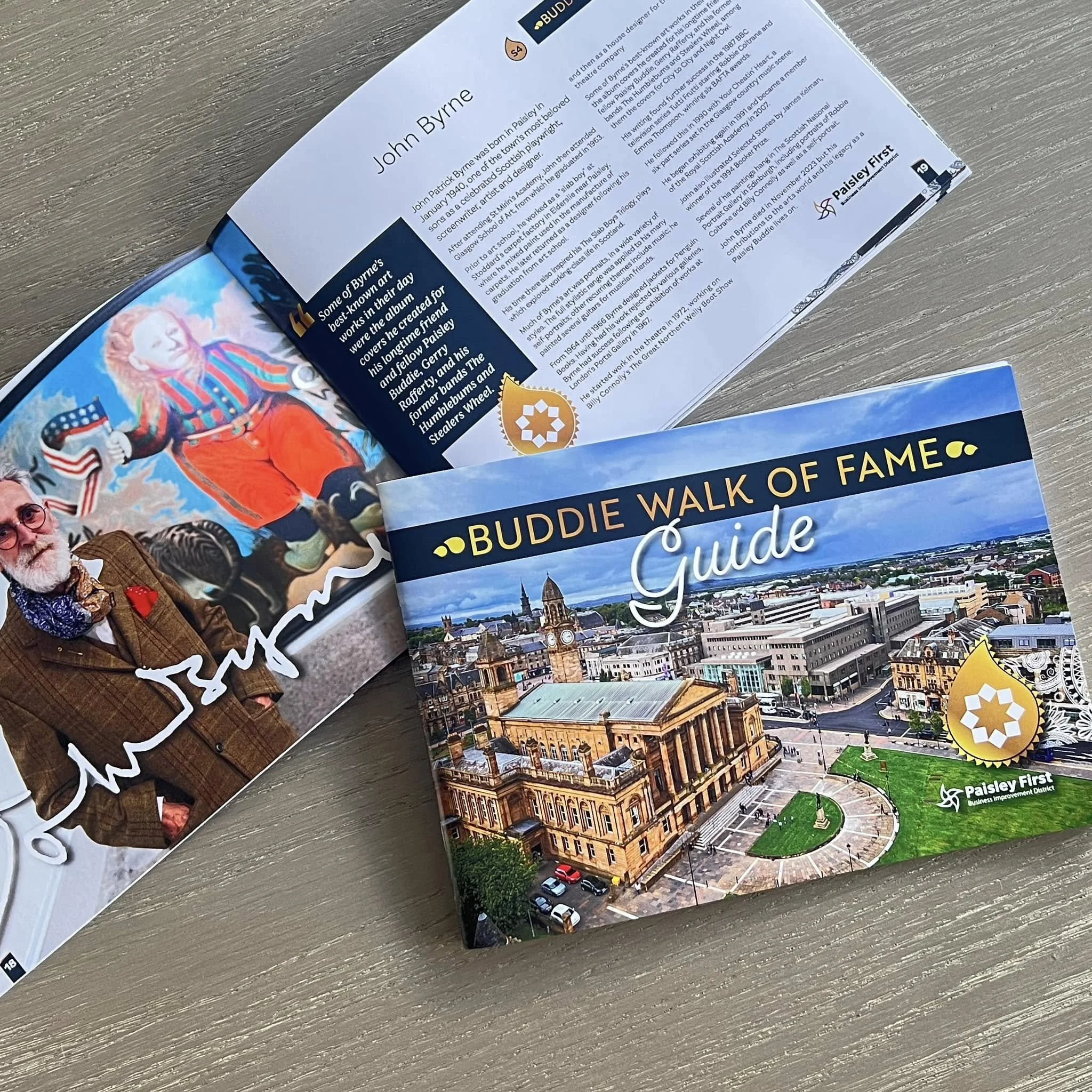 Cover of a Buddy Walk of Fame guidebook featuring an aerial view of a city, with historic buildings and a clock tower, and a sticker with a star logo. The inside reveals a biography of John Byrne with a photograph of him in a blazer and a signature.