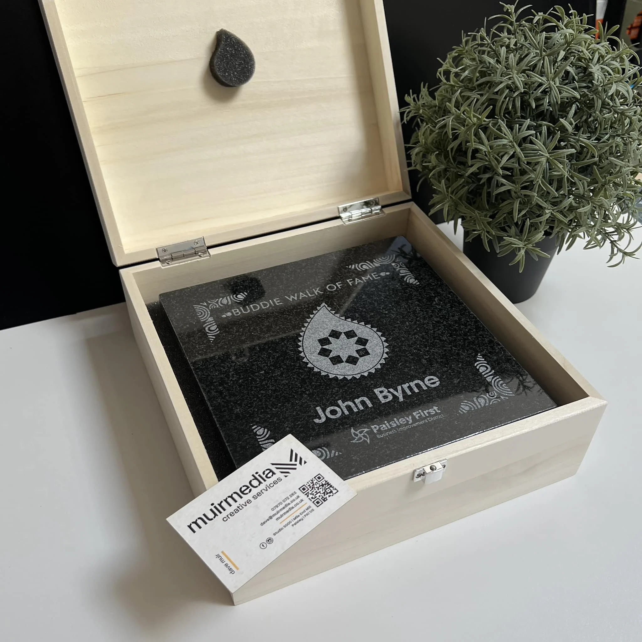 Wooden box containing a black engraved plaque with the text 'BUDDIE WALK OF FAME,' a droplet shape, and the name 'John Byrne.' Next to the box is a potted green plant.