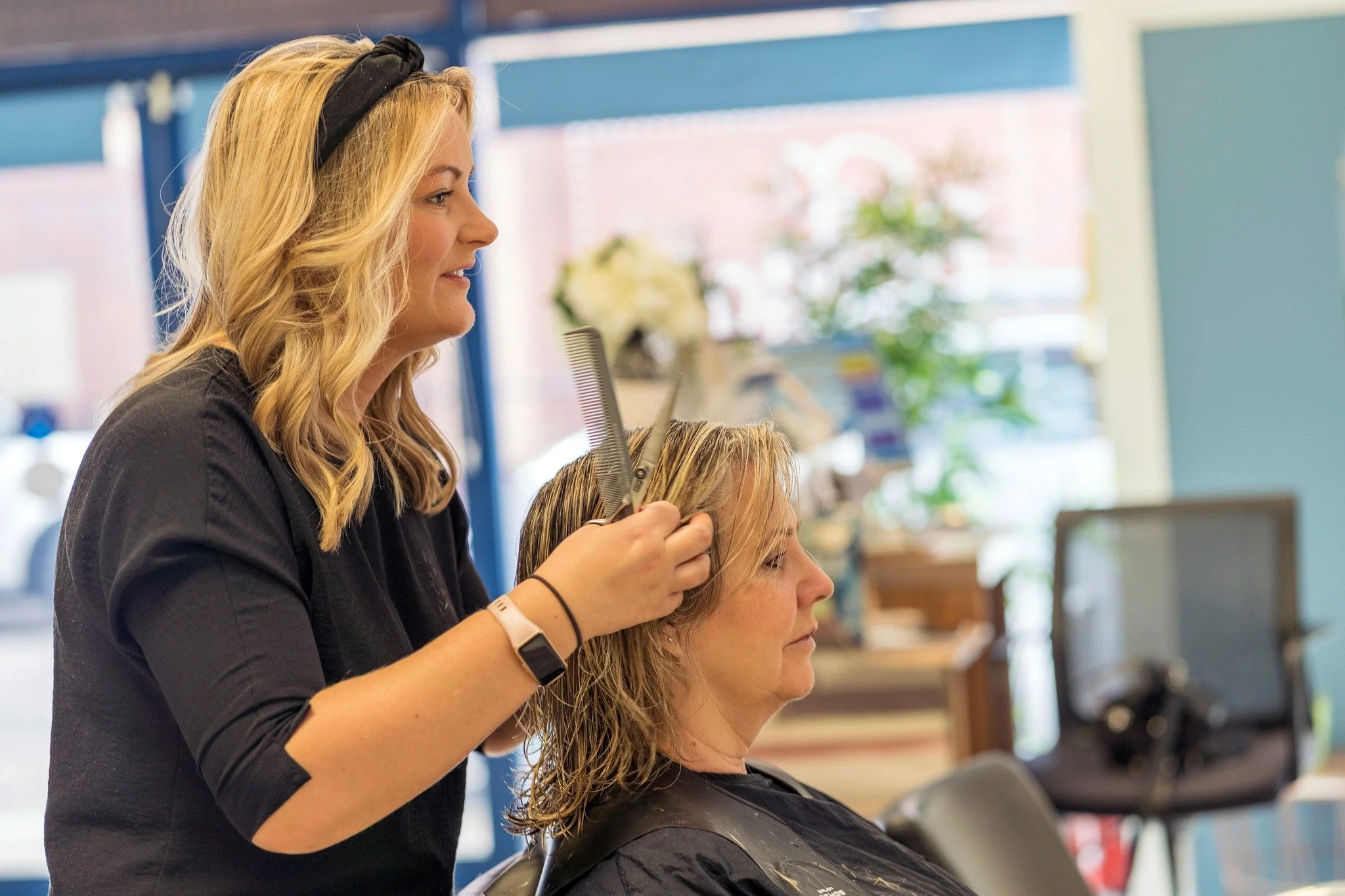A hairstylist trims a woman's hair in a salon with bright natural light and a colorful background.