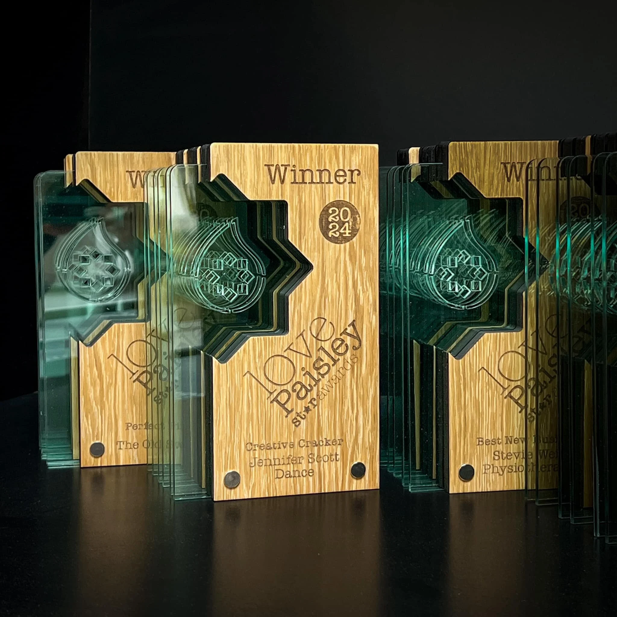 Set of glass awards with wooden plaques, inscribed with titles and names, including one labeled 'Winner 2024' in gold color, standing on a black surface with a dark background.