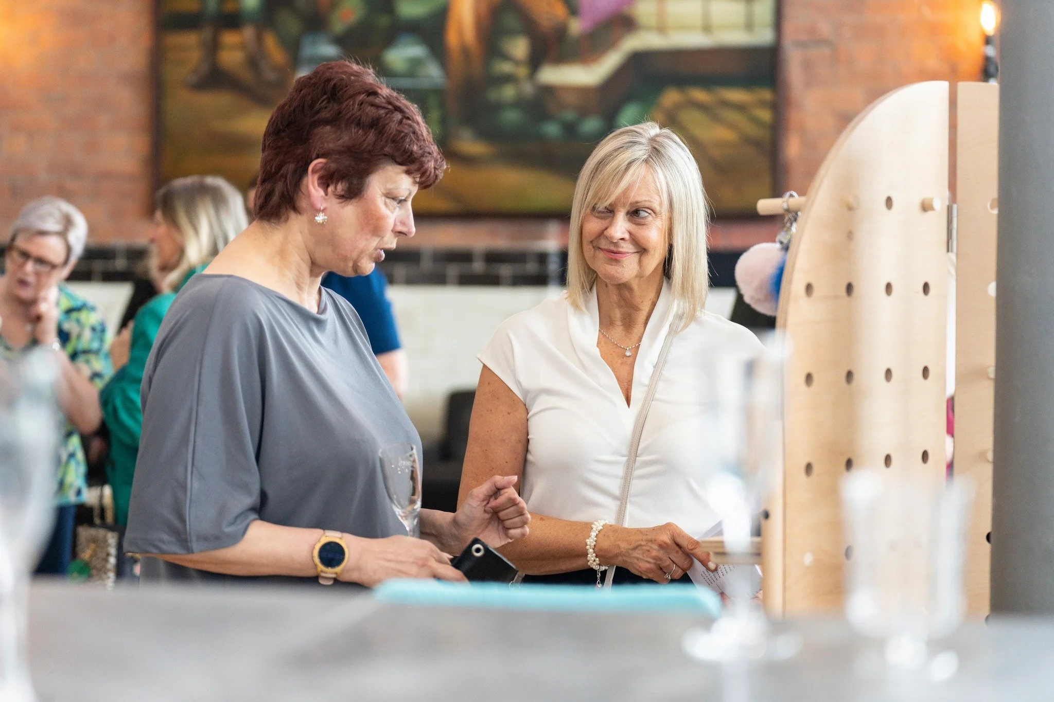 Two women are standing and talking at a social event or gathering. There are other people in the background, with a brick wall and artwork on the wall.