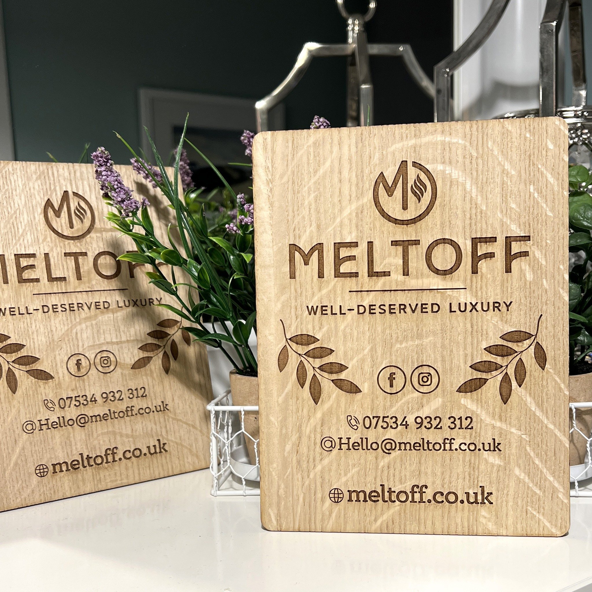Wooden business cards for Meltoff, a luxury brand, with contact info and social media icons, placed next to purple flowering plants on a white surface.