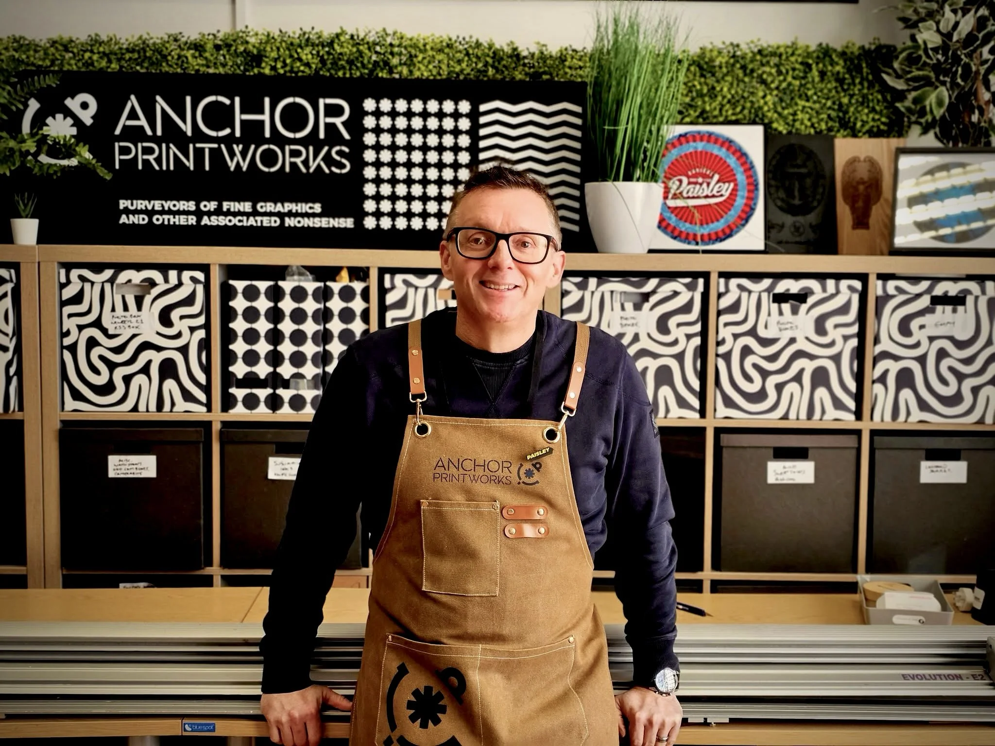 A man with glasses and a black sweatshirt wearing a tan apron standing in front of display shelves with patterned boxes, a sign reading 'Anchor Printworks', and various artwork, inside a store or print shop.