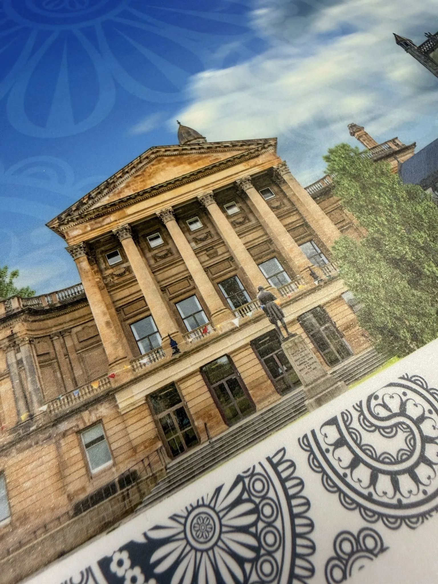 Photo of a neoclassical building with large columns, a triangular pediment, and a statue on top, seen through a reflective surface with design patterns and a blue sky in the background.