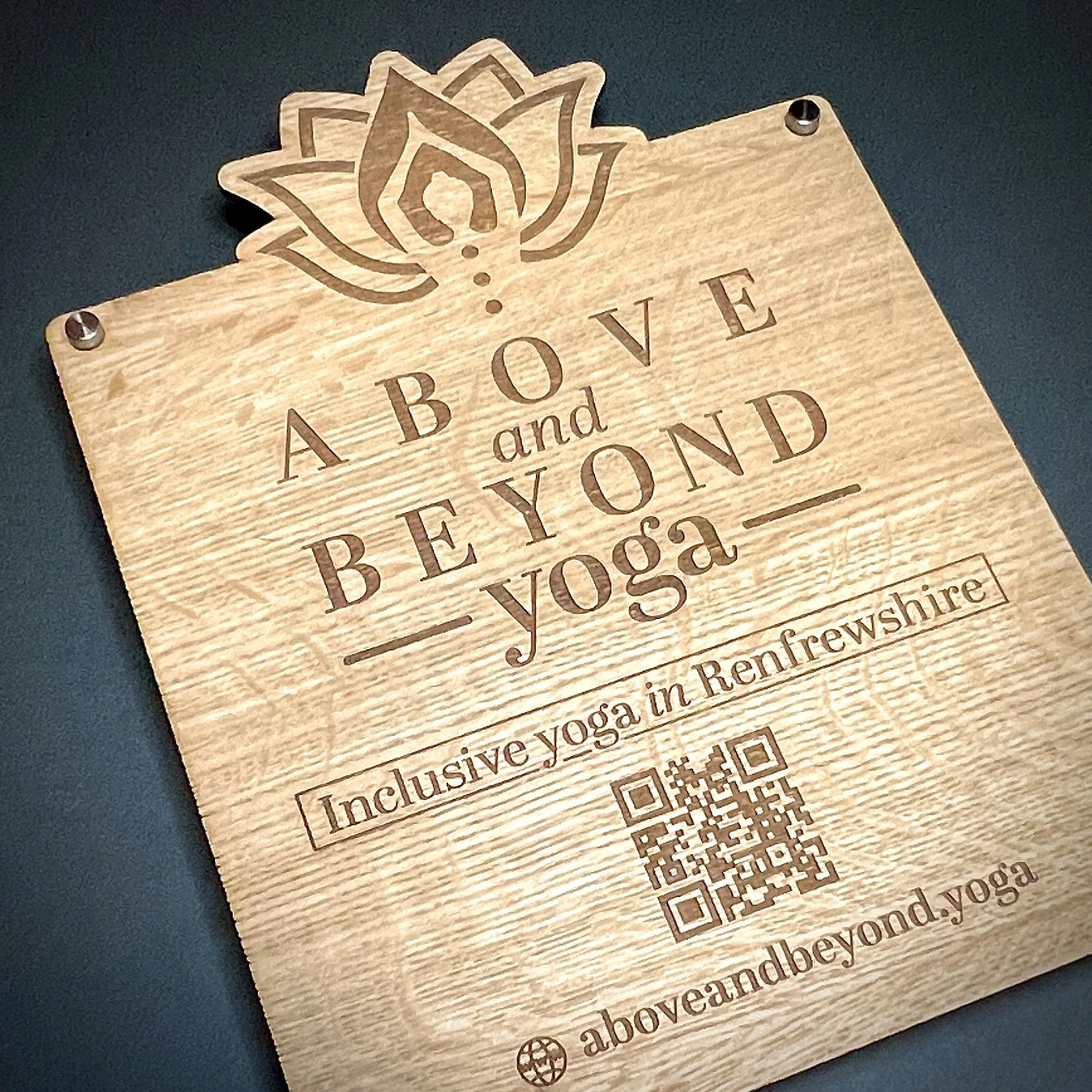 Wooden sign with lotus flower design, advertising inclusive yoga in Renfrewshire, Scotland, featuring a QR code and website URL.
