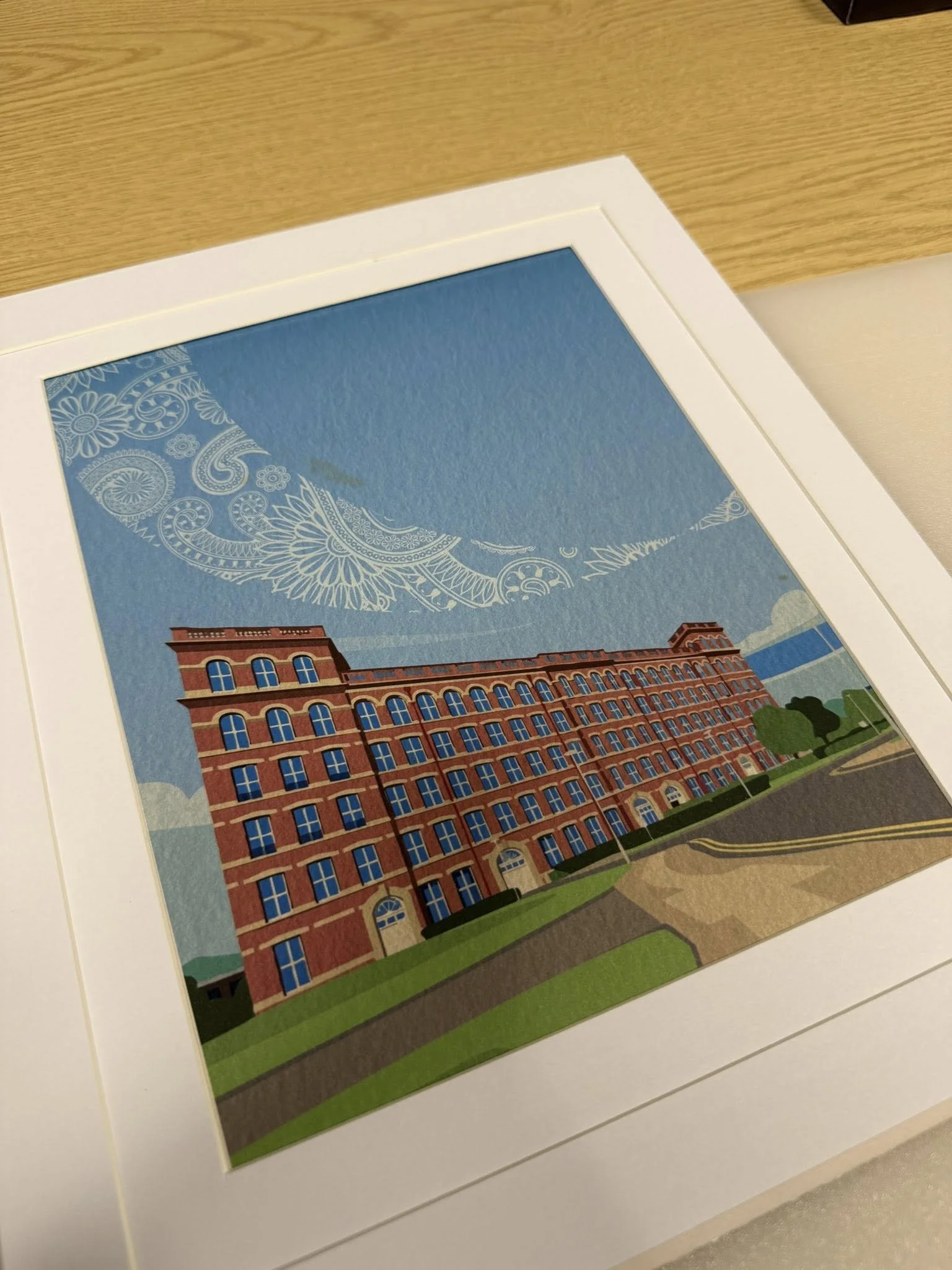 Colorful framed illustration of a historic brick building with multiple windows, set against a sky with decorative patterns and clouds, on a table.