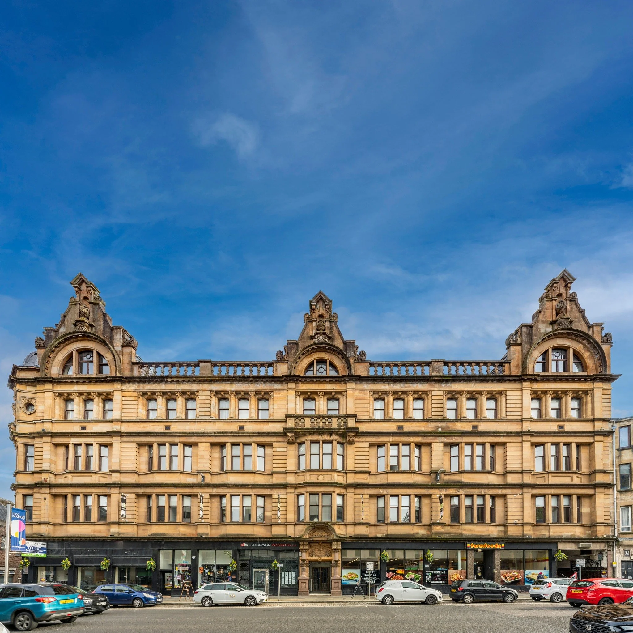 A historic multi-story building with classical architecture, featuring ornate details and a dark storefront on the ground floor, parking and cars in front, under a blue sky.