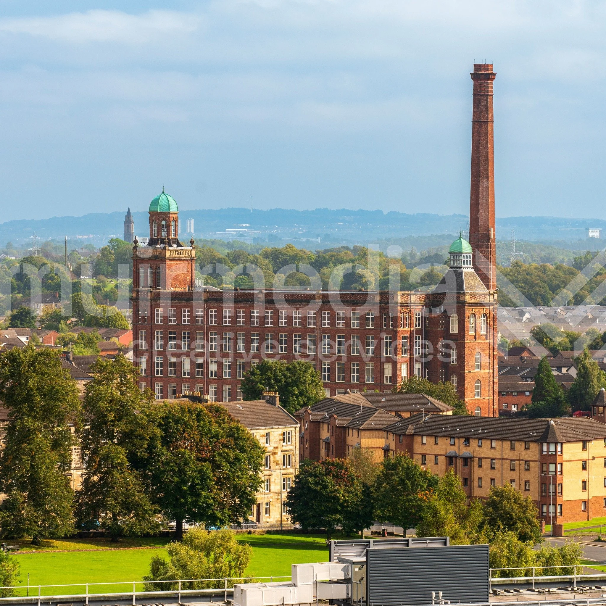 View of a historic red-brick building with green domes and a tall chimney, surrounded by trees, residential buildings, and distant hills.