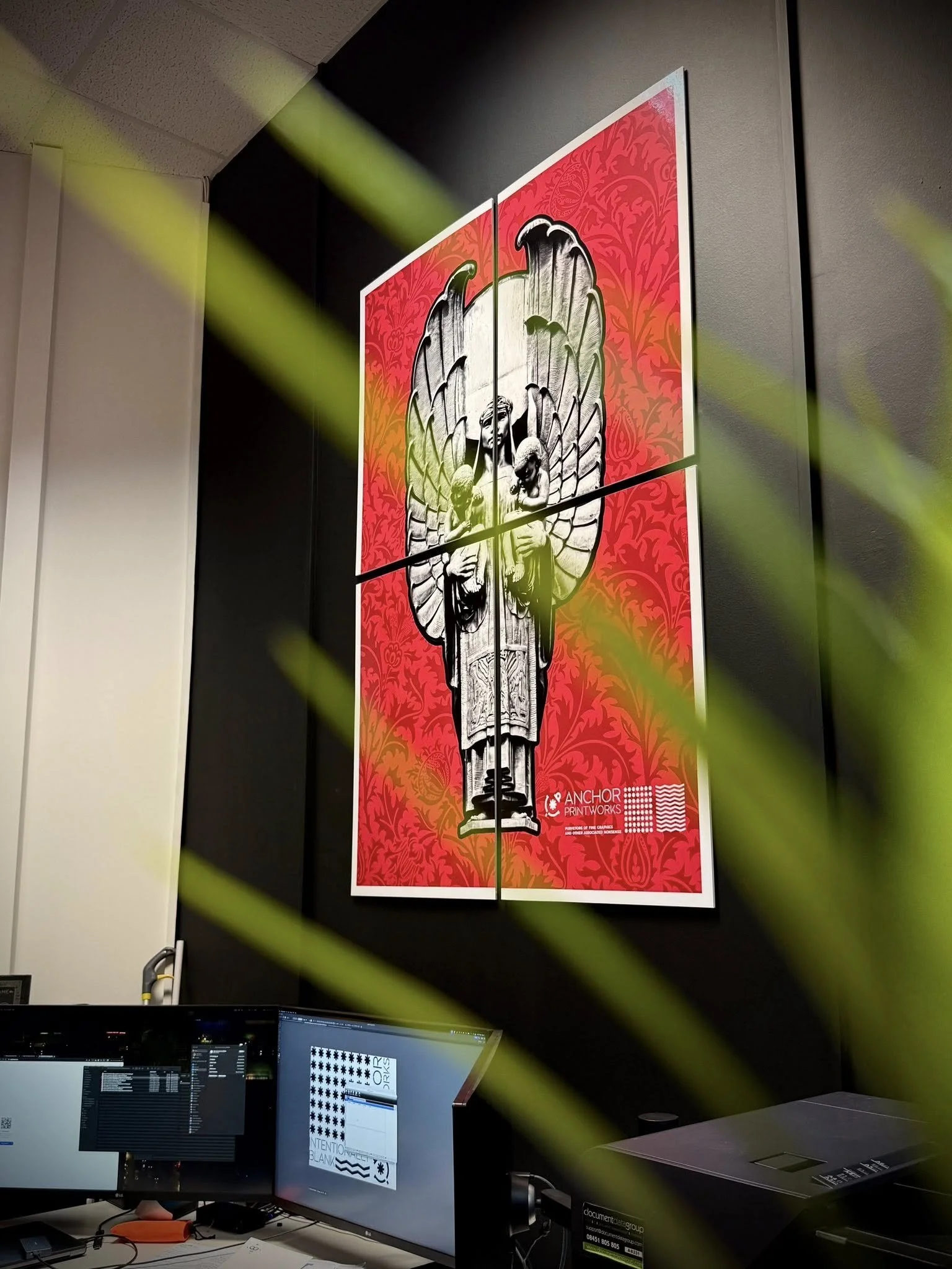 A framed artwork on a black wall depicting a stylized image of a religious or saintly figure with wings, against a vibrant red and orange patterned background, located in an office or workspace with computer monitors and equipment in the foreground.