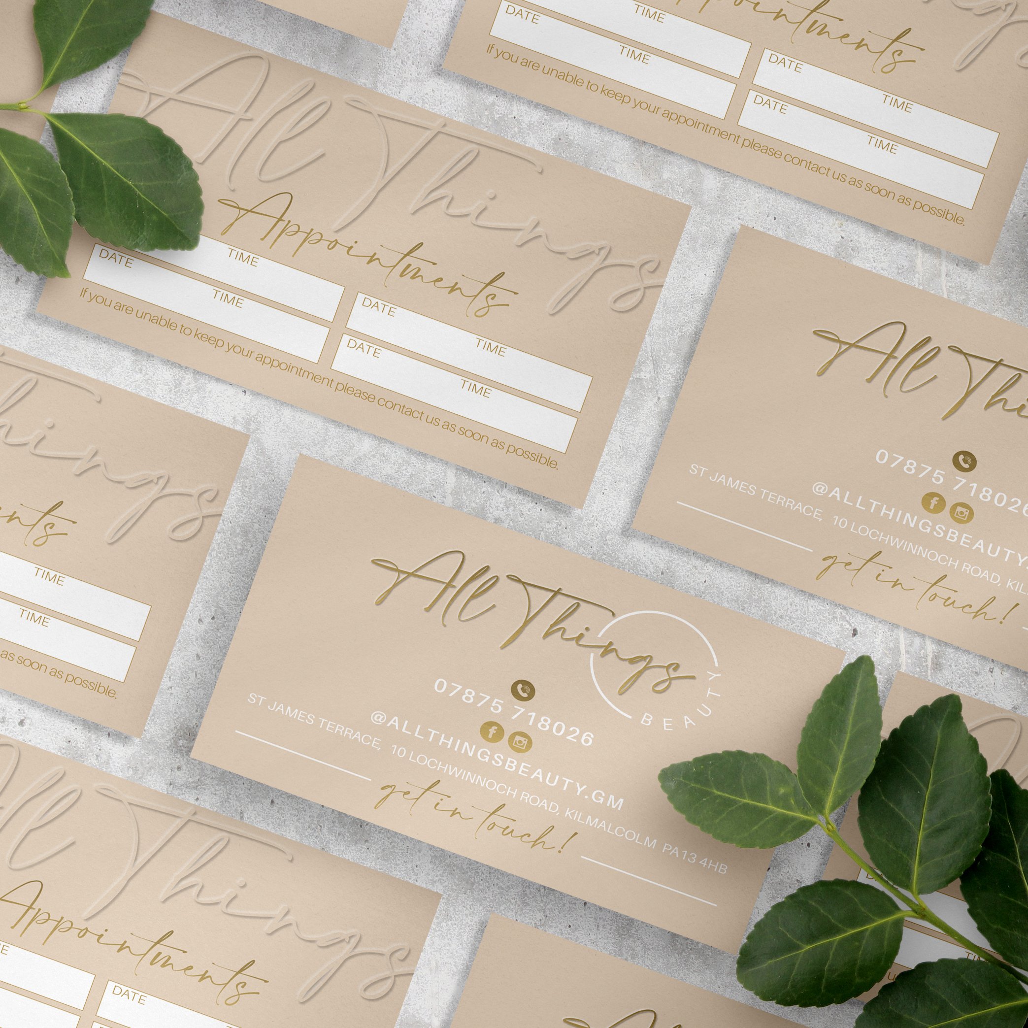 Business cards for All Things Beauty with contact information, social media icons, and appointment scheduling details, laid out on a textured surface with green leaves.
