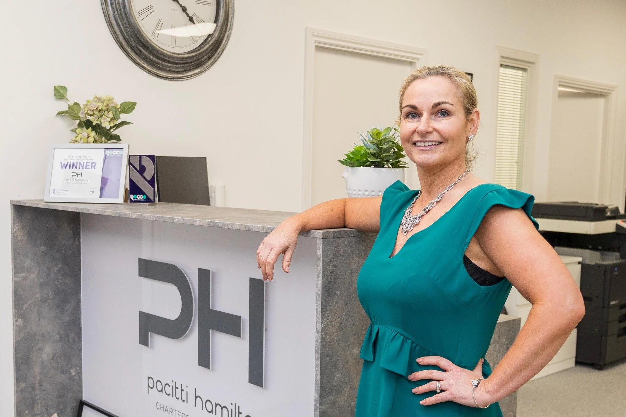 A smiling woman in a teal dress stands at a reception desk with her arm resting on it, in an office environment. Behind her is a sign that reads 'PH paciti hamilton,' a potted plant, a framed certificate, and a clock on the wall.