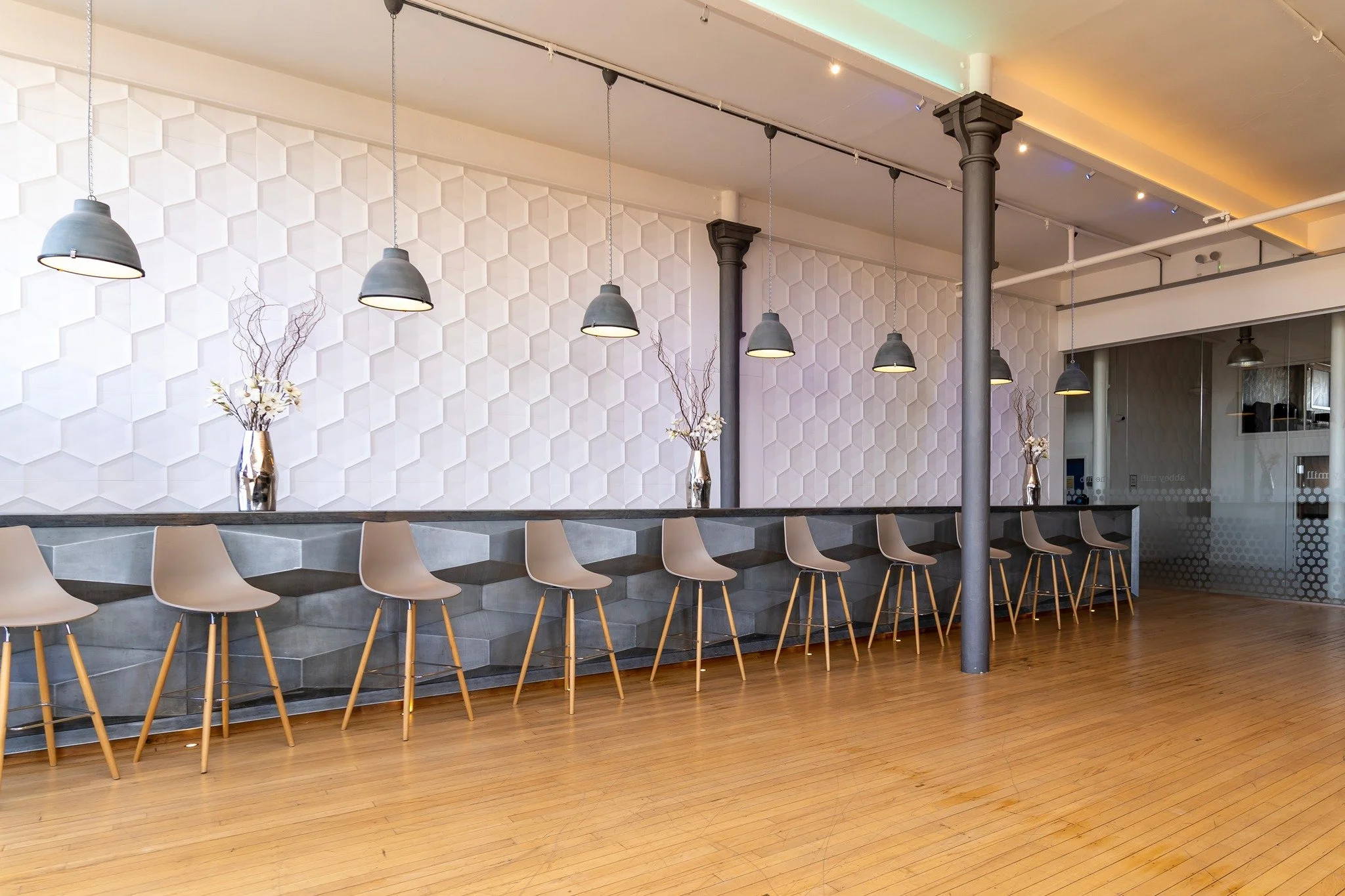 Modern cafe interior with a row of bar stools along a high counter, decorated with vases and floral arrangements, pendant lighting, wood flooring, geometric wall tiles, and a glass wall/door in the background.