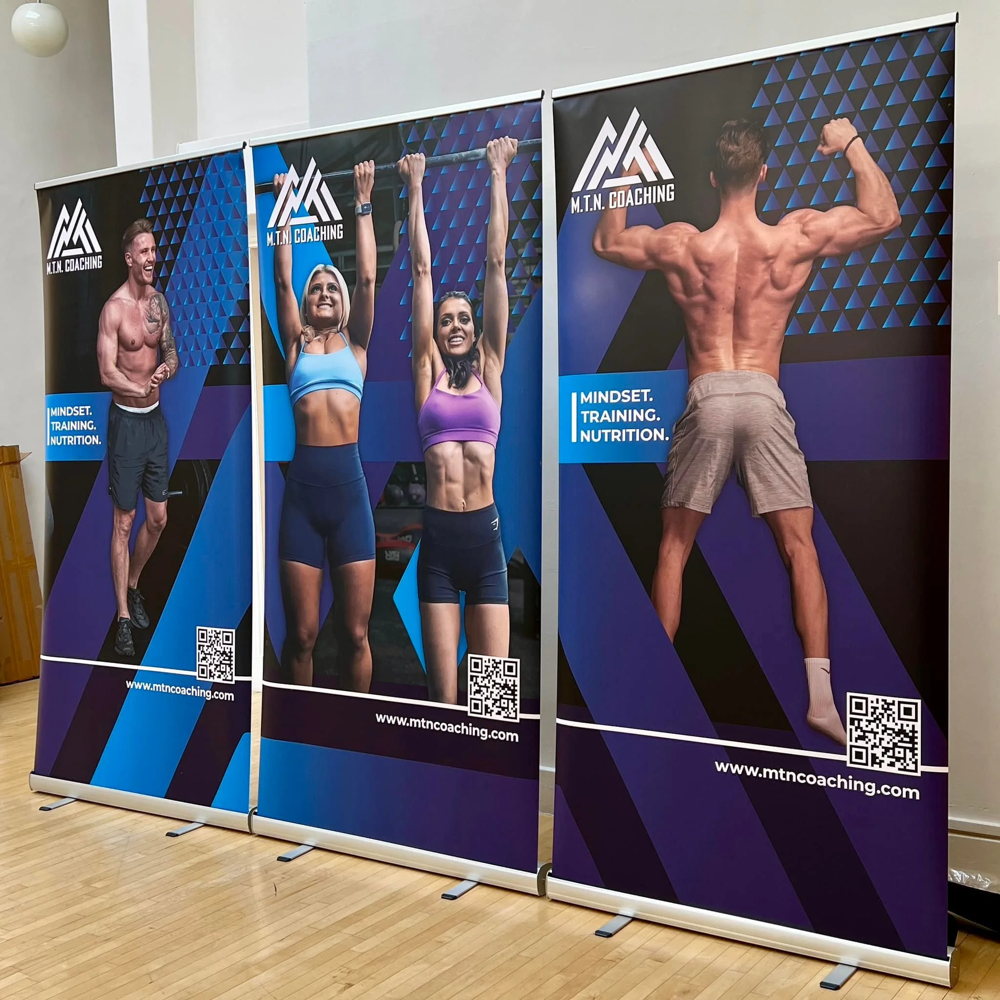 Three large fitness posters display muscular and fit individuals engaging in different workouts. The first poster features a shirtless man with tattoos, the second shows two women in athletic clothing hanging from a pull-up bar, and the third depicts