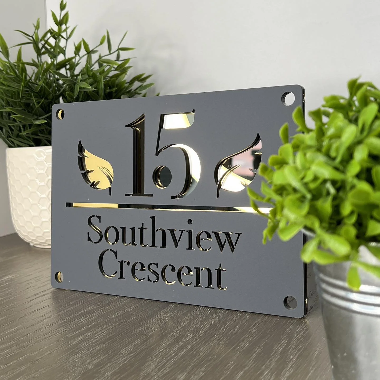 A metal house number sign displaying '15' and 'Southview Crescent' surrounded by potted plants on a wooden surface.