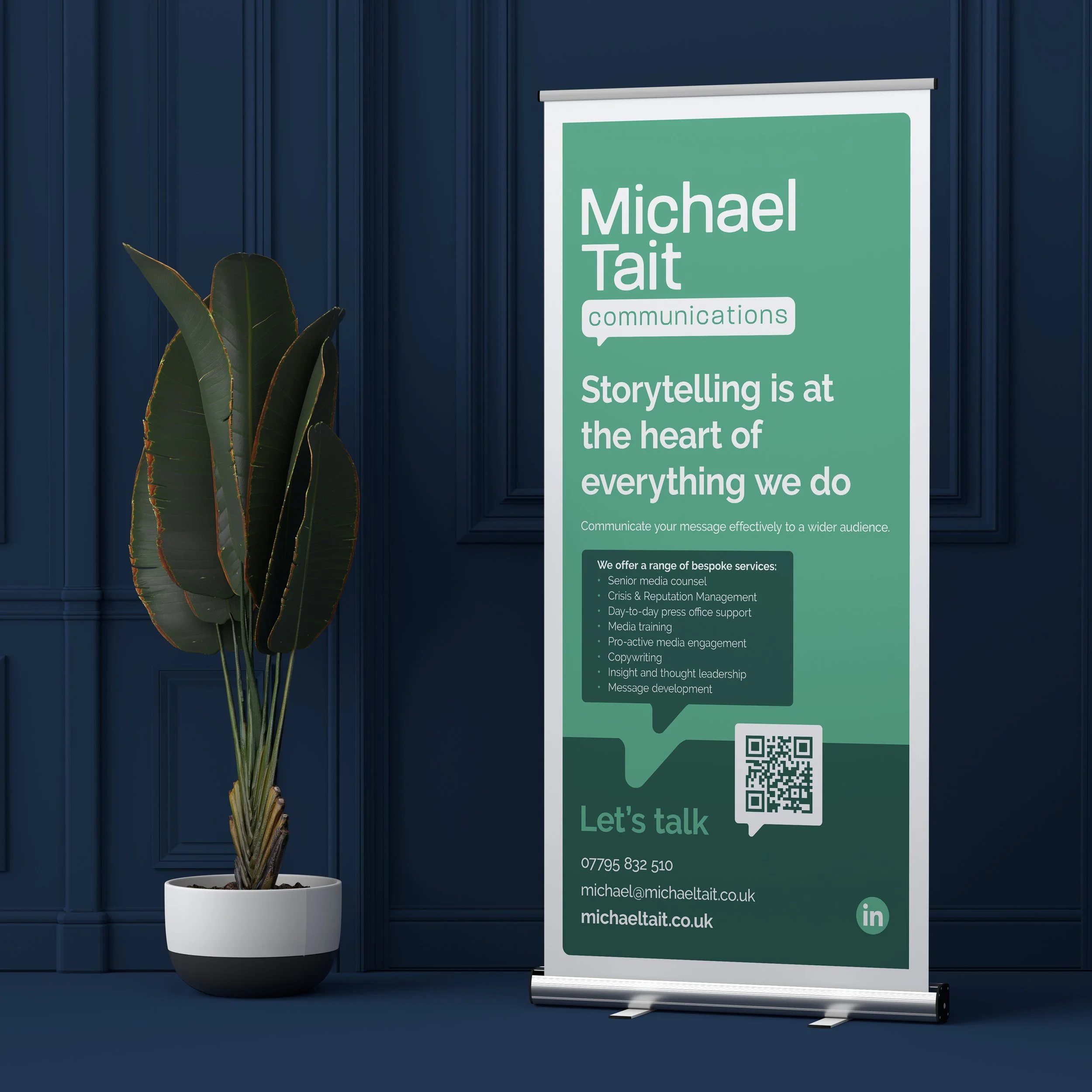 A vertical banner standing next to a potted plant with large green leaves. The banner is green and white with text advertising communication services by Michael Tait, emphasizing storytelling. It includes a QR code, contact information, and a LinkedI