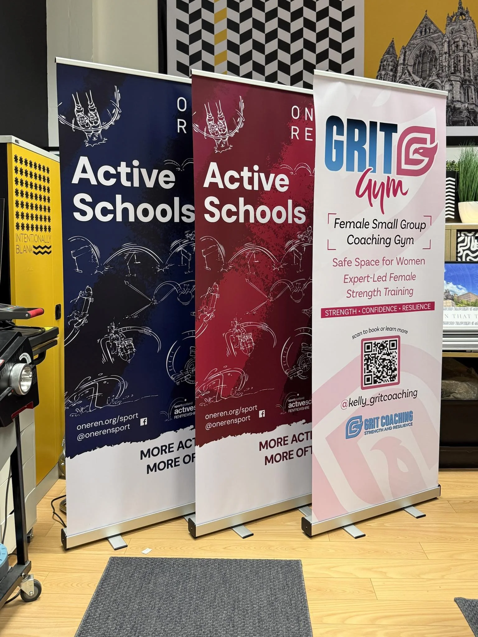 Three vertical banners promoting active sports and a female coaching gym, placed on a wooden floor with some wall art in the background. The first two banners advertise 'Active Schools,' one in blue and one in red, with website and social media info.